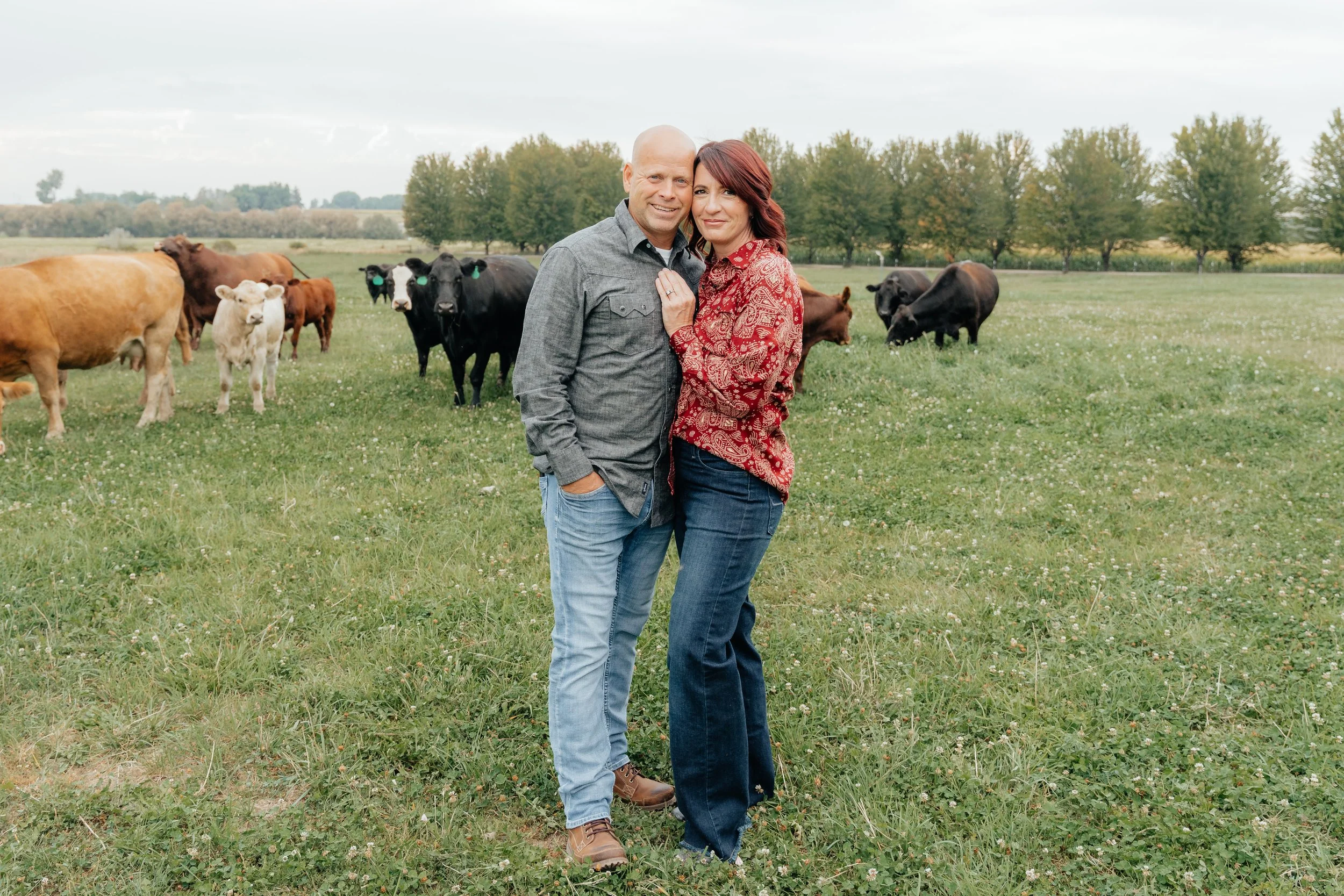 Boise Doodle Co.'s owners! A couple standing close together in a field of grass with cows grazing behind them, trees in the background, under a partly cloudy sky.