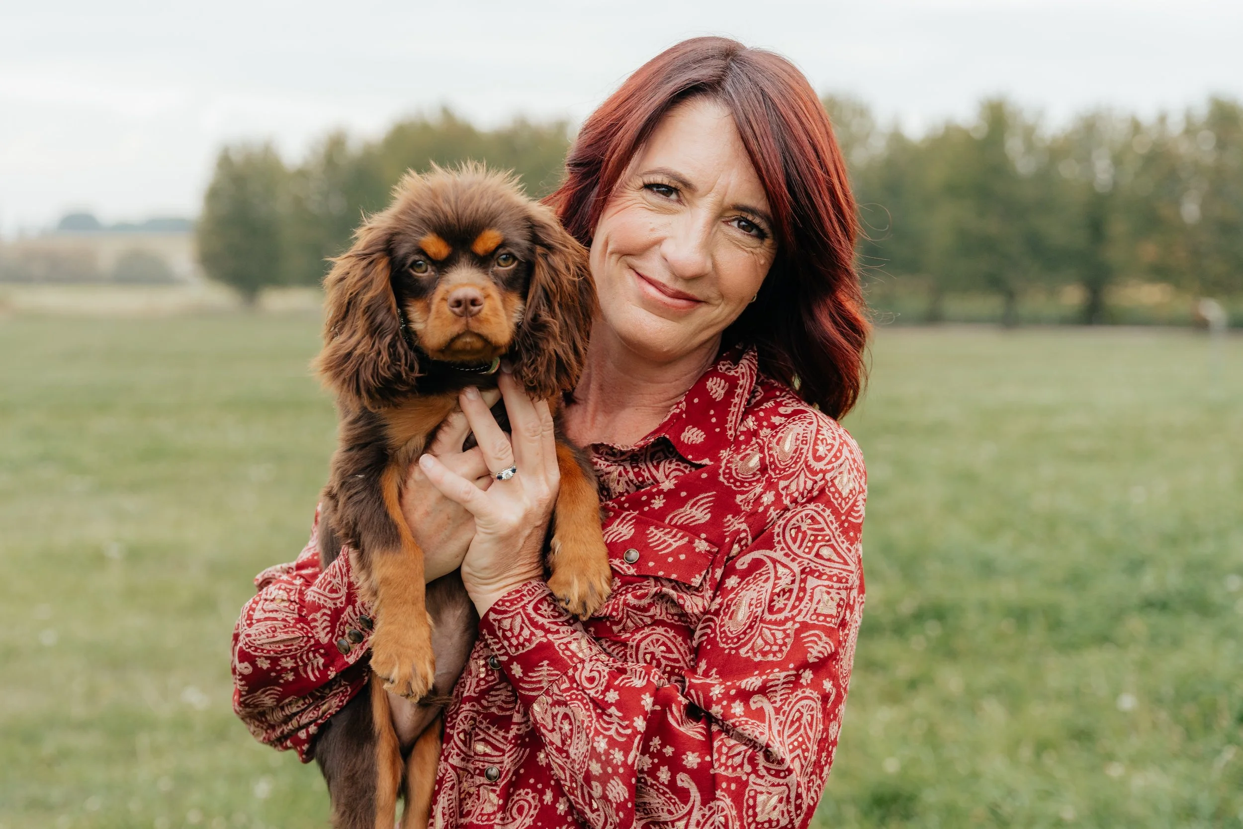 A woman with red hair holding a brown and black puppy outdoors in a green field with trees in the background.