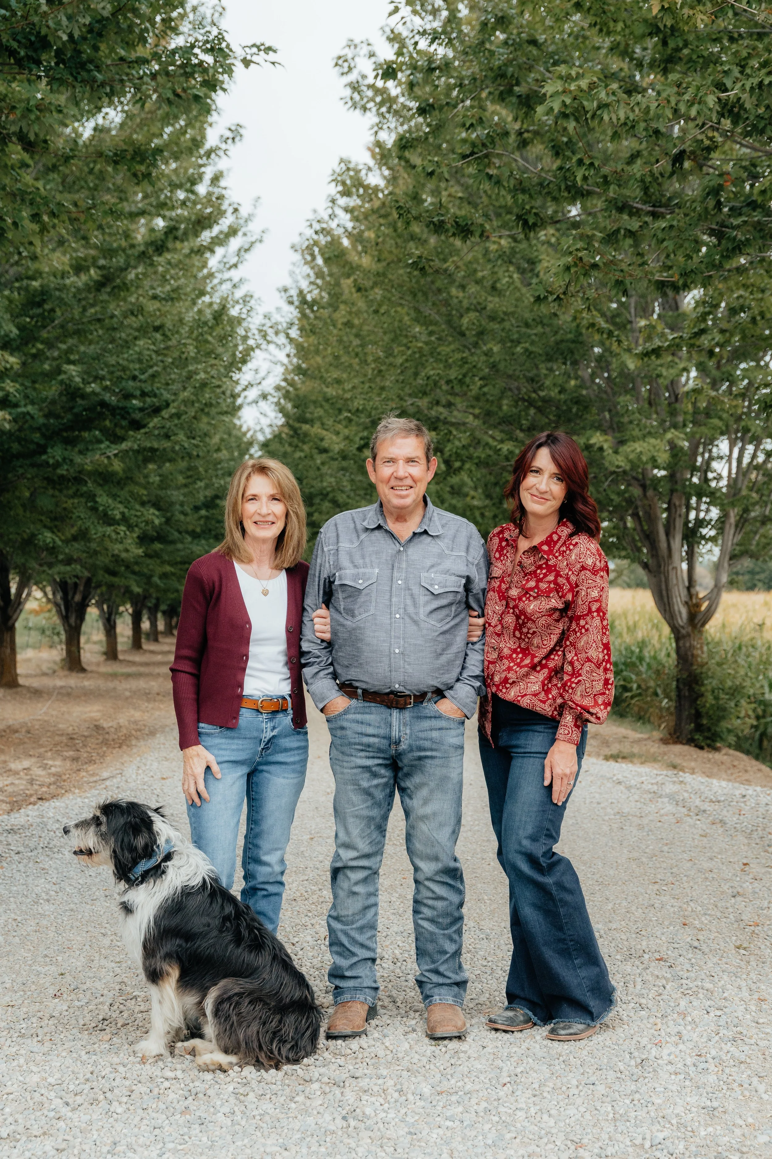 Group of three adults and a dog in a park with trees in the background. The woman on the left has blonde hair and wears a burgundy cardigan, white shirt, and blue jeans. The man in the middle has short graying hair and wears a gray button-up shirt and jeans. The woman on the right has dark red hair and wears a red patterned blouse and jeans. The dog is a black and white Border Collie sitting on the gravel path in front of them.