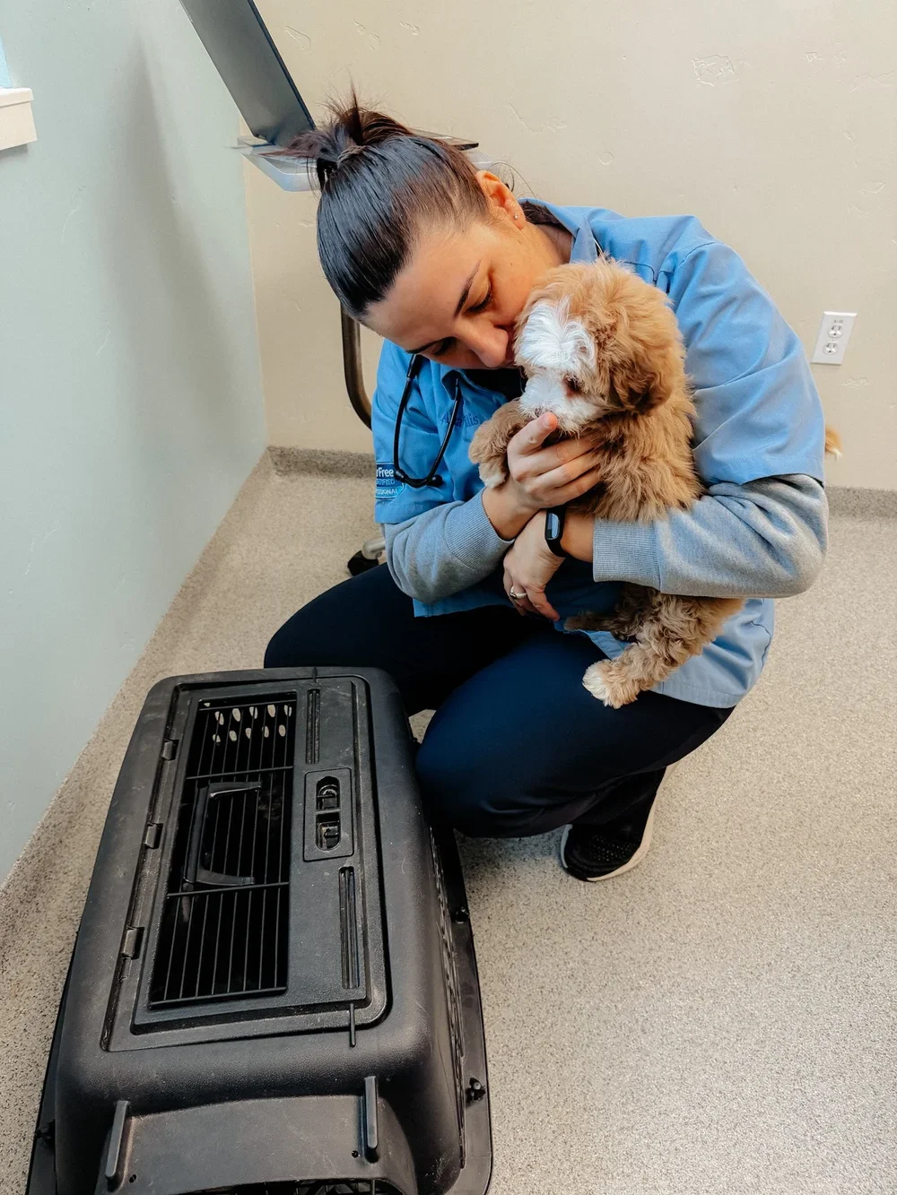 A woman in scrubs, kneeling on the floor, holding a small, fluffy brown and white puppy close to her face in a veterinary clinic or animal hospital. There is a portable air conditioner or heater on the floor in front of her.
