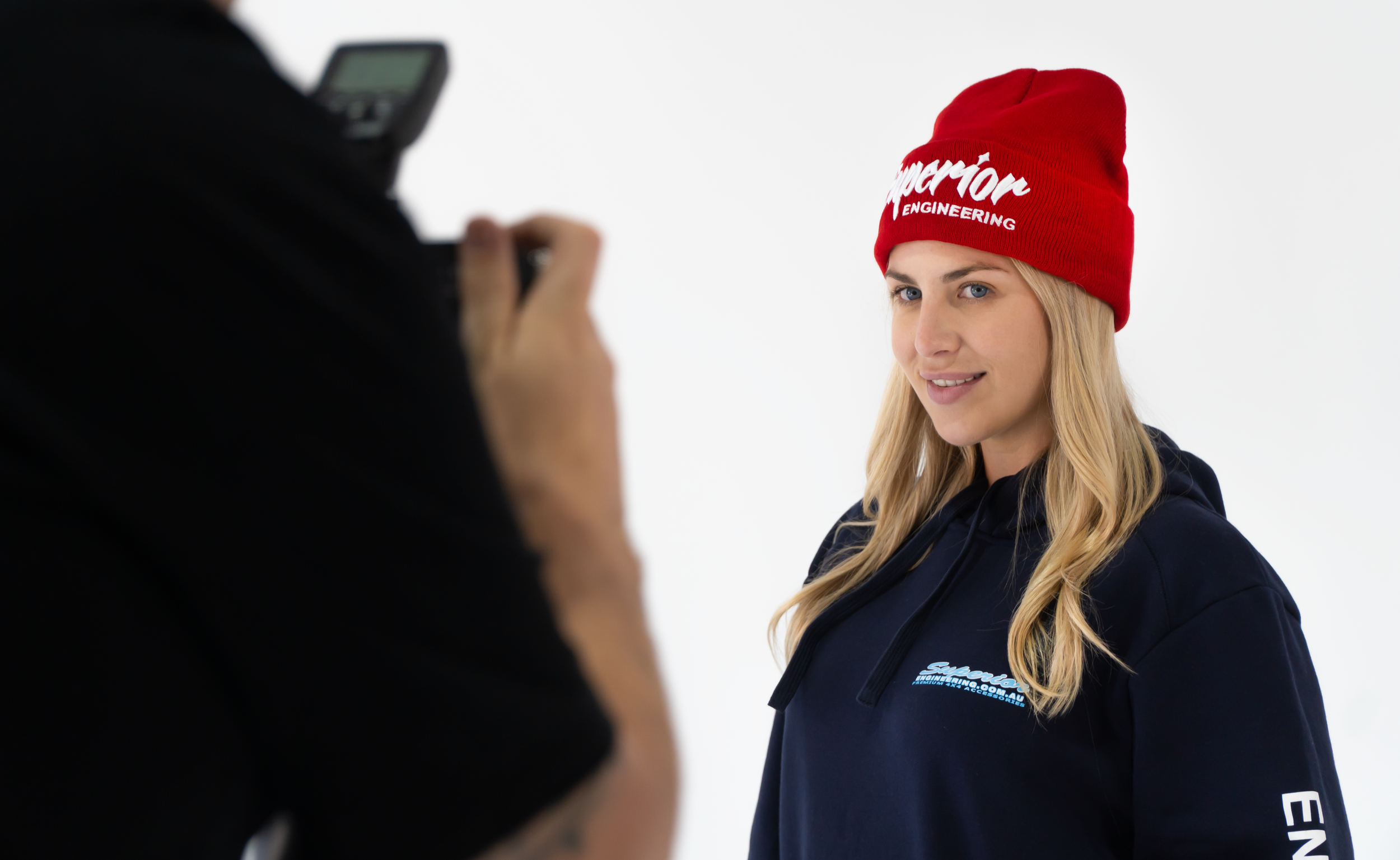 A woman in a red beanie with white lettering and a navy jacket is being photographed or filmed against a plain white background.
