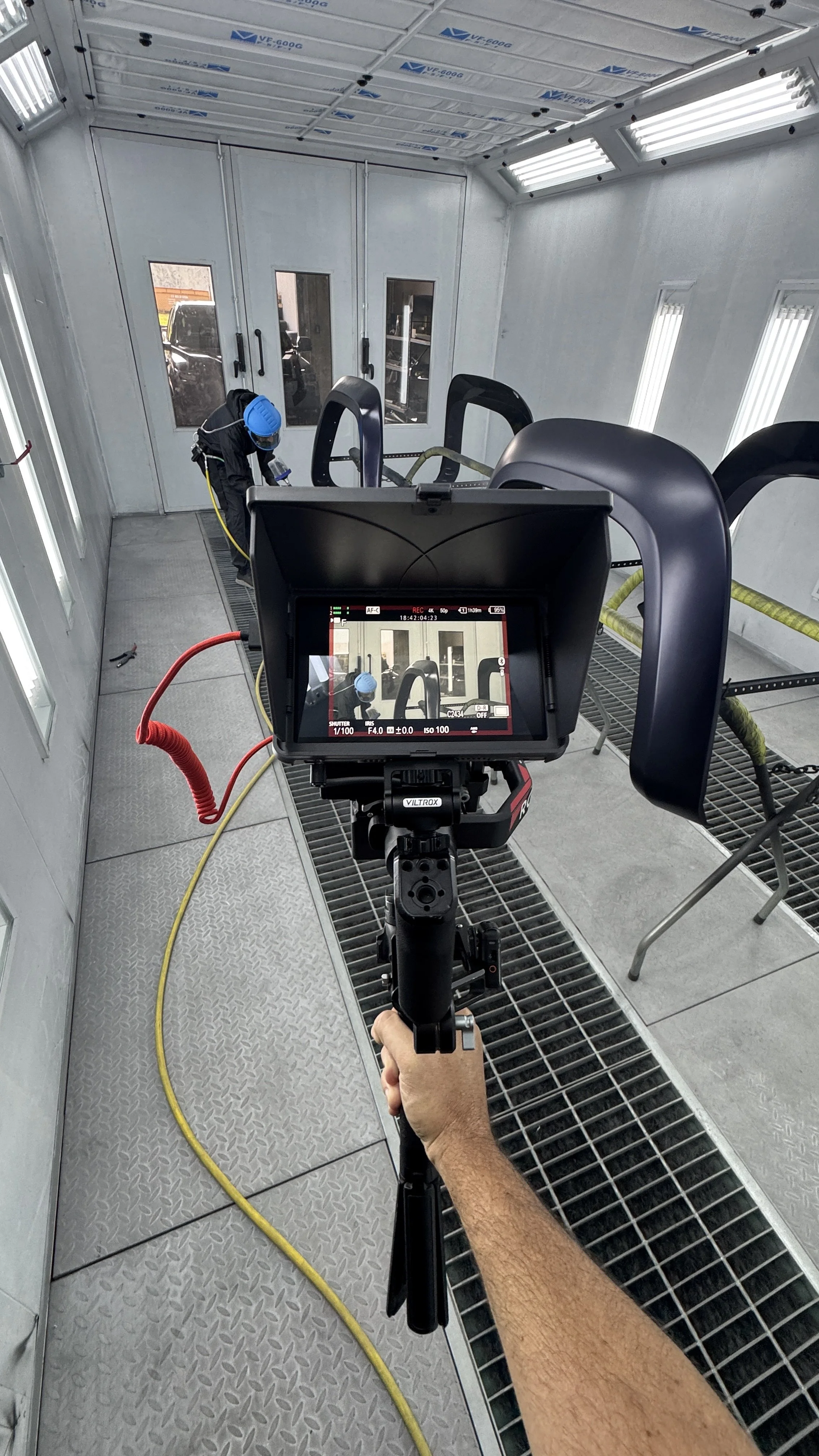 Inside a spray booth with a person wearing protective gear working on a vehicle part, and a camera on a tripod recording the process.
