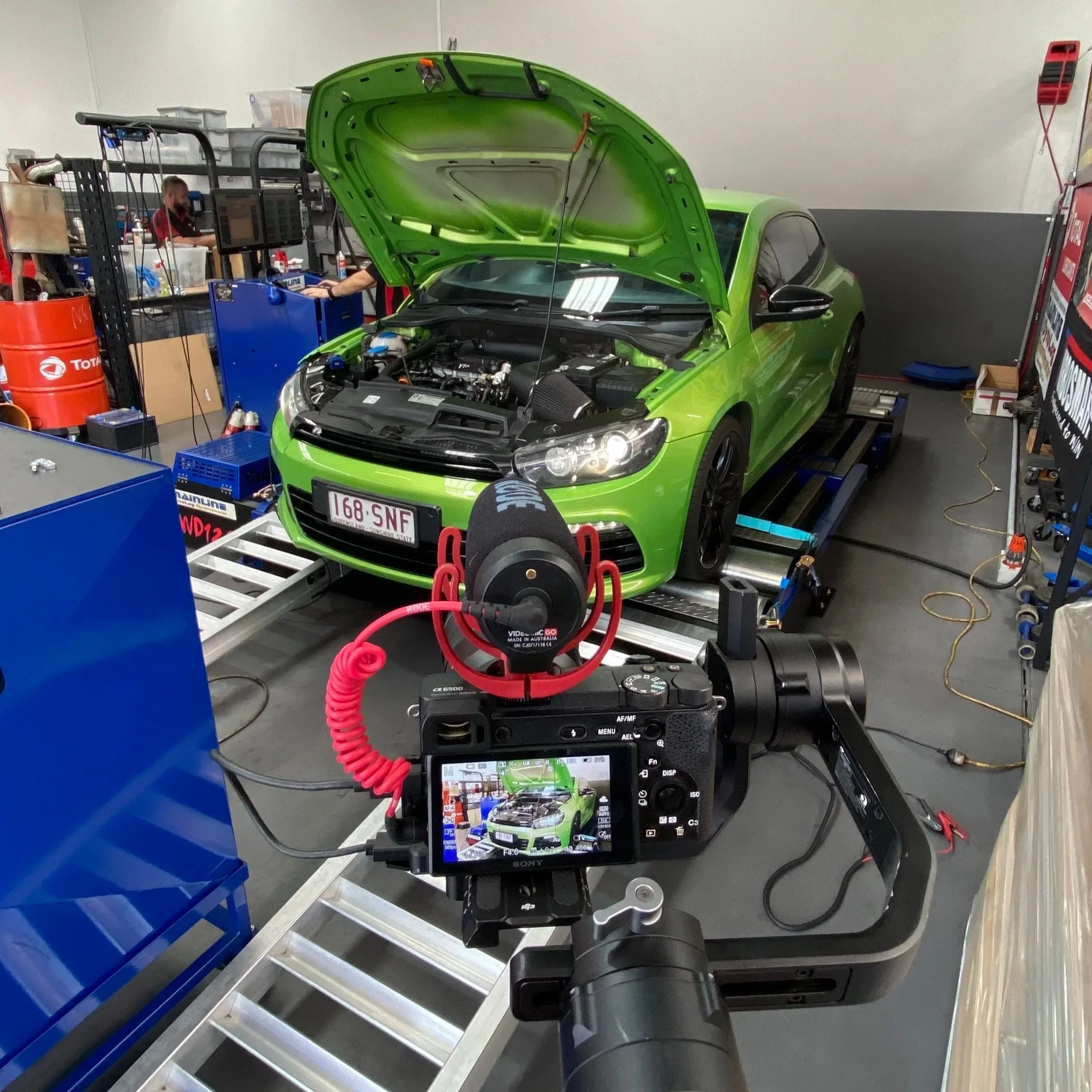 A bright green car with its hood open is on a dynamometer in a garage or workshop. A camera and other equipment are positioned; a person is working in the background at a workstation with tools and containers.