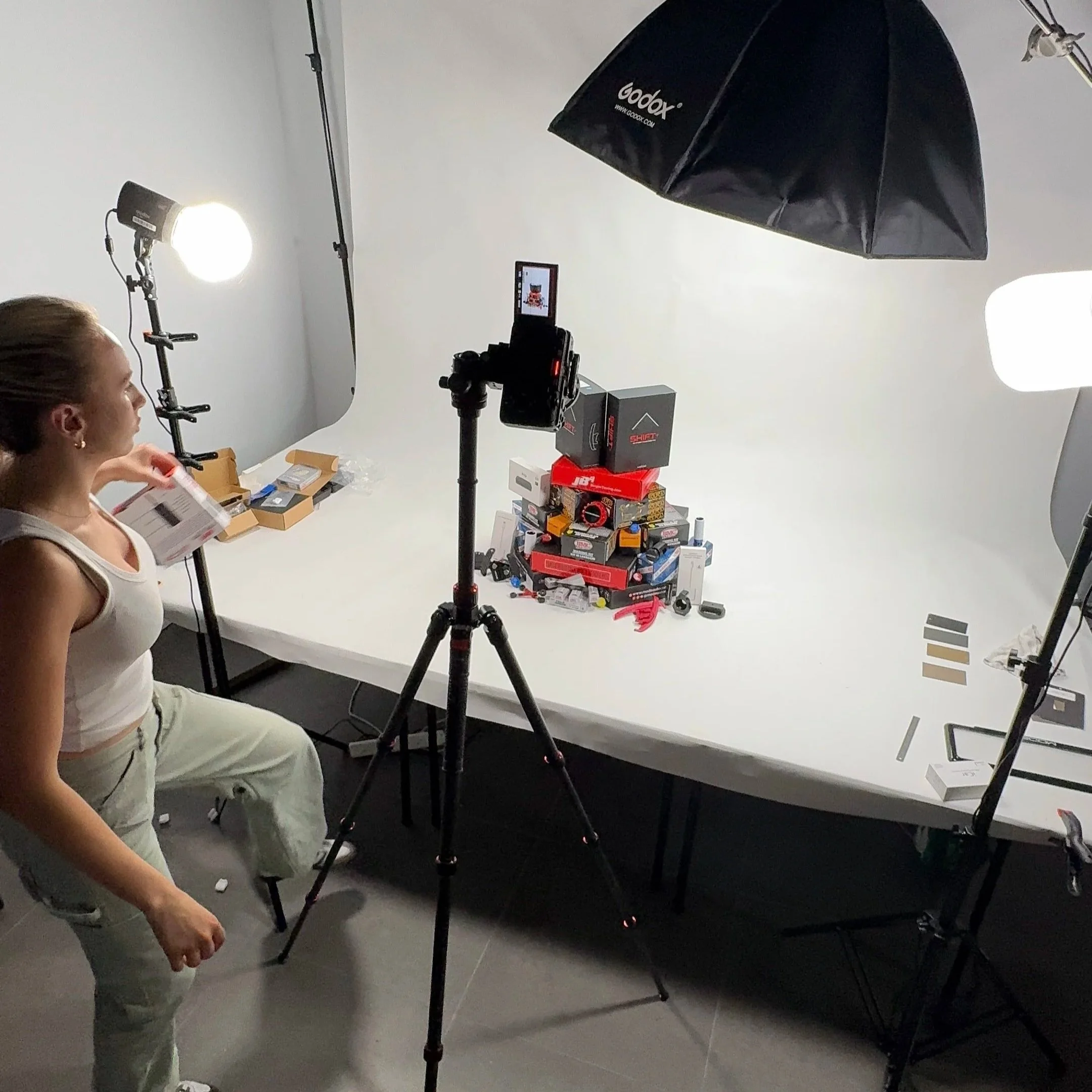A woman is sitting in a photography studio setup with professional lighting and equipment, including a large umbrella reflector and a camera on a tripod. She is holding a box and appears to be in the process of an activity or shoot, with shelves of b