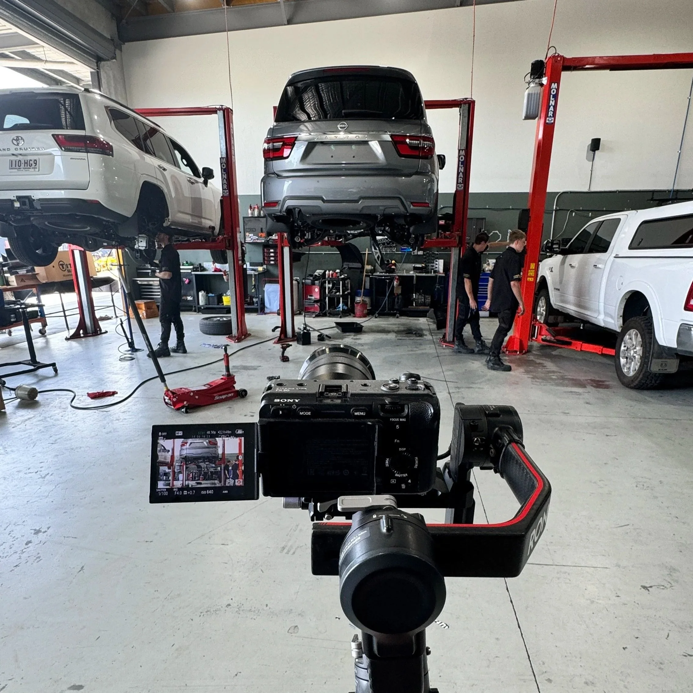 Inside an auto repair shop, a vehicle is elevated on a hydraulic lift, with several mechanics working around it. There are tools, equipment, and other vehicles visible in the background.