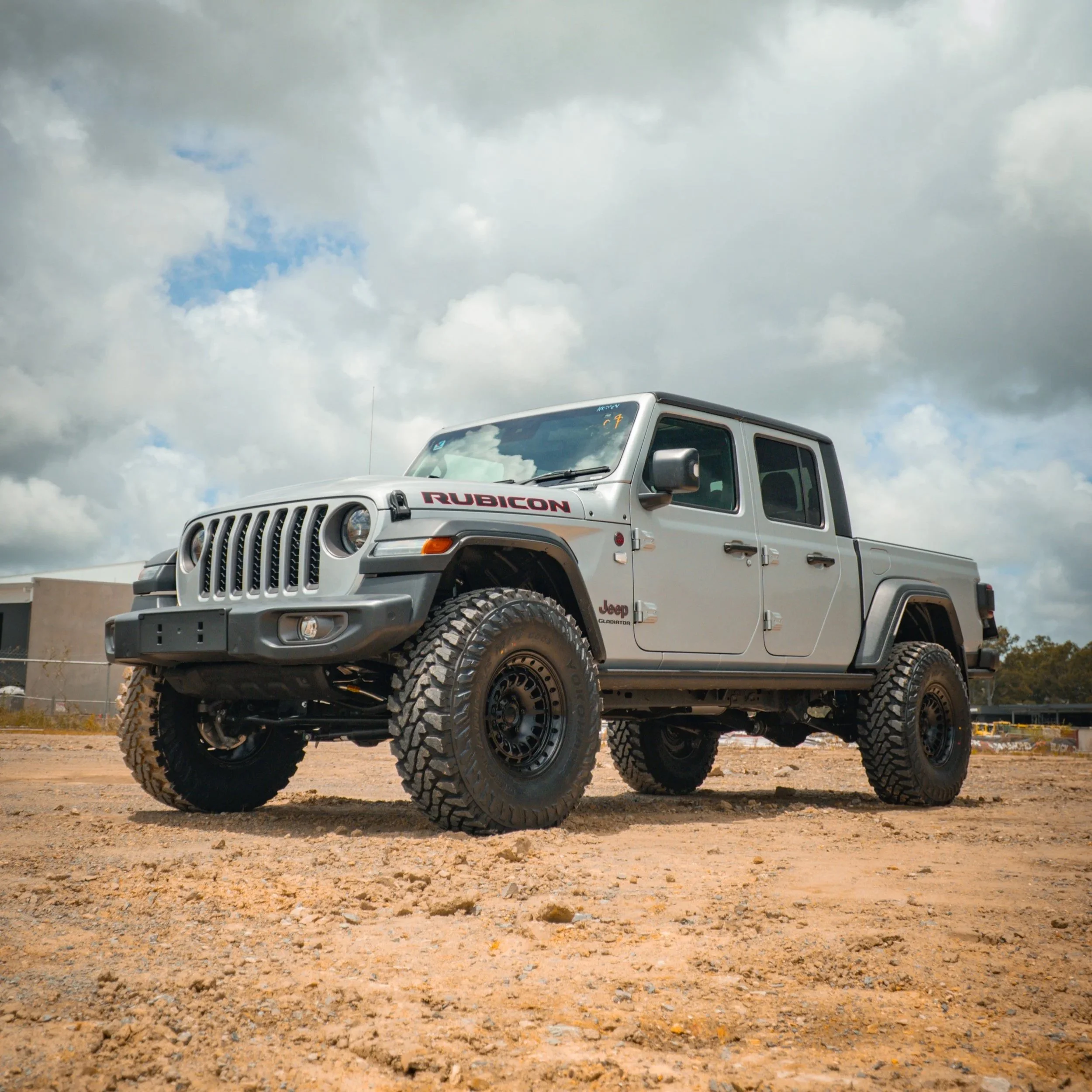 A silver Jeep Rubicon Gladiator pickup truck parked on a dirt lot under a cloudy sky.