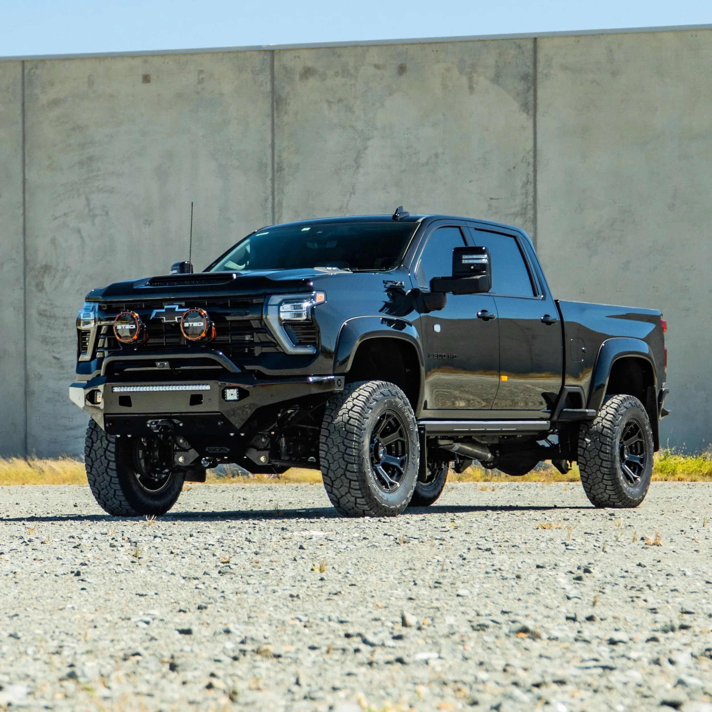 A black Chevrolet Silverado truck with off-road tires, a front bumper with integrated LED lights, and a hood scoop, parked on a gravel surface in front of a plain beige wall under a clear blue sky.