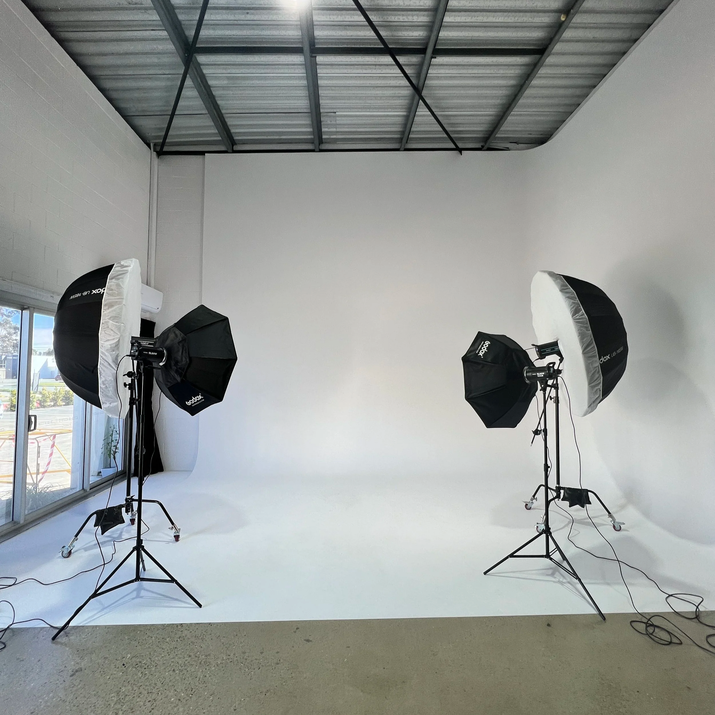 Photography studio setup with multiple softbox lights on adjustable stands arranged in front of a white seamless backdrop in a well-lit room with large windows.