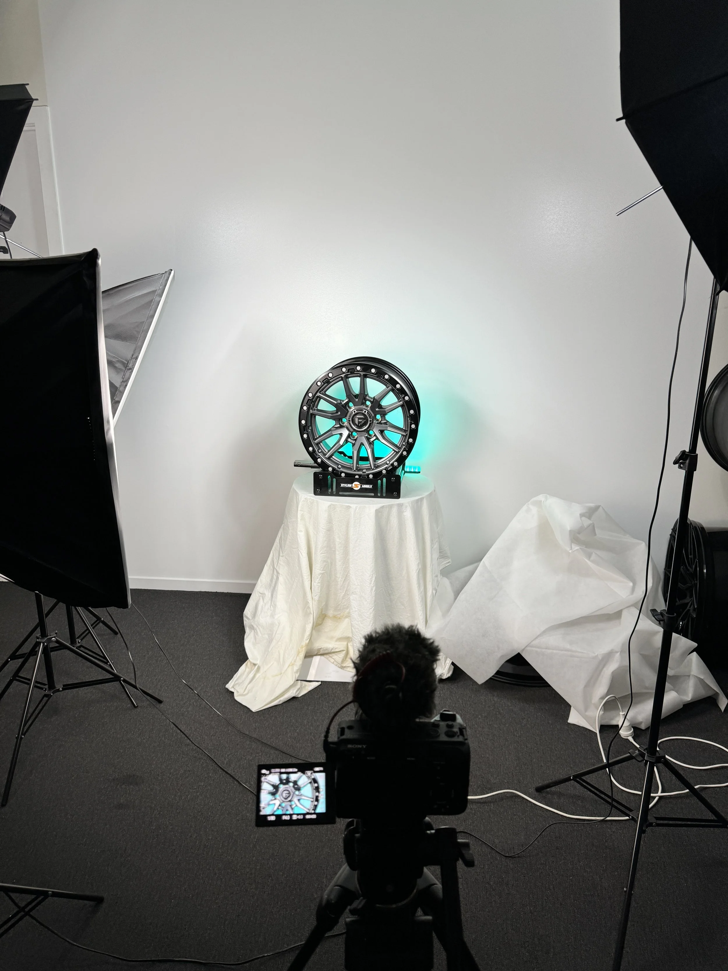 A photography studio setup with studio lights and a white backdrop. In the center, a wheel rim is placed on a small table covered with a white cloth, and a camera is capturing the shot in the foreground.