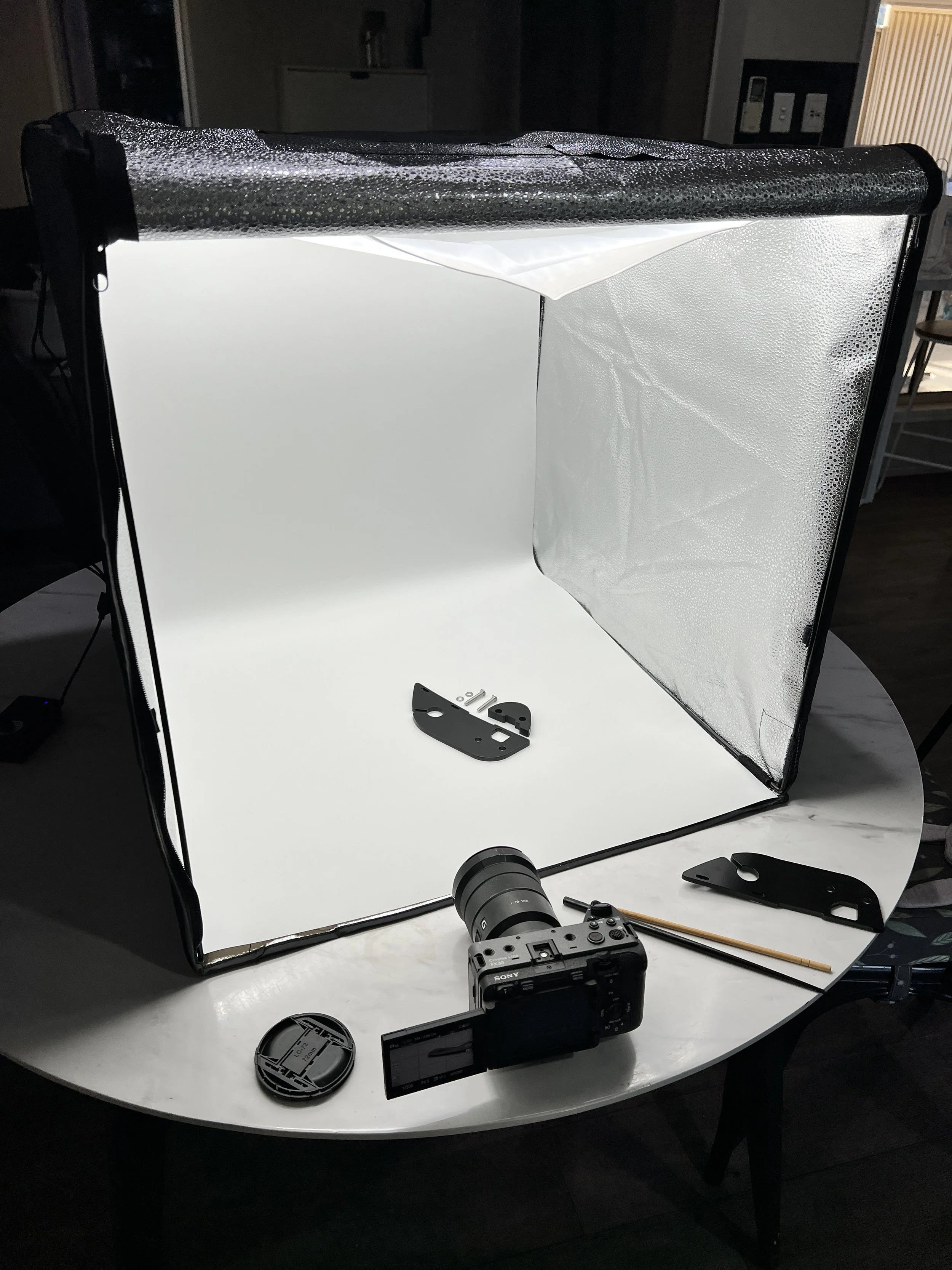 A photography setup with a white backdrop, a reflective silver reflector, camera equipment, and small accessories on a white table, used for professional photo shoots.