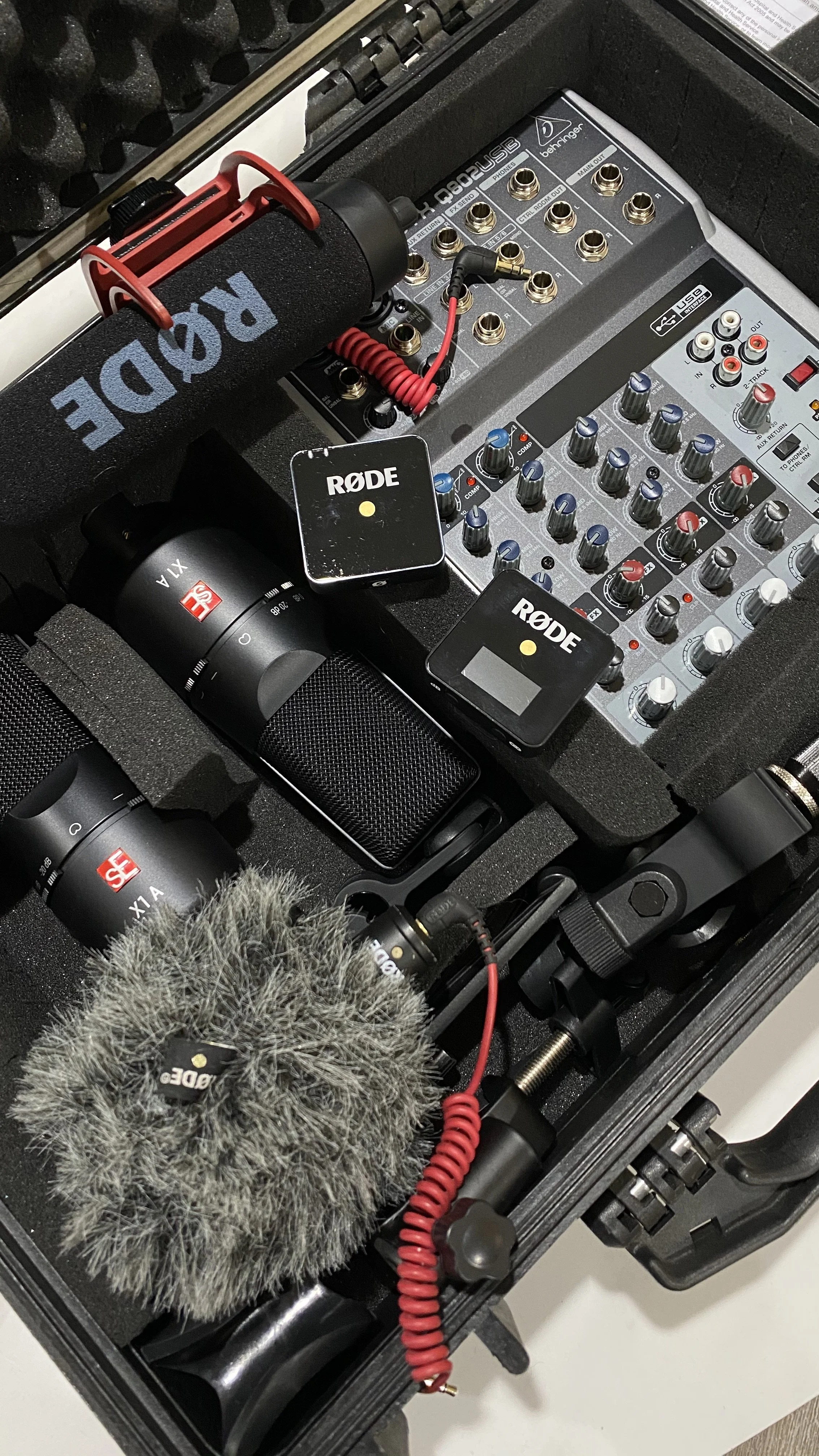 Audio recording equipment inside a case, including a microphone with a red shock mount, audio interfaces, a large furry windscreen, and various cables and connectors.