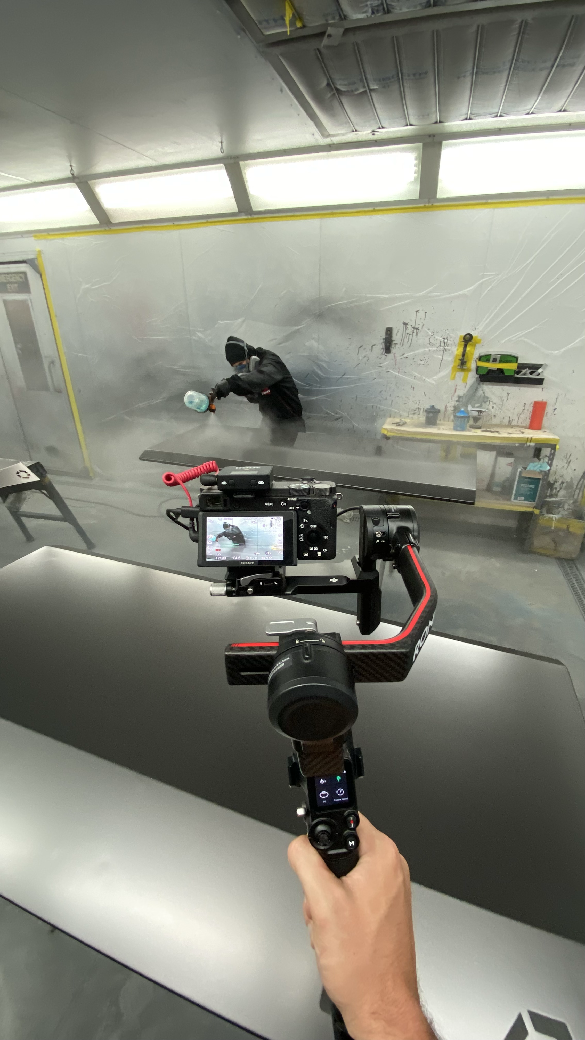 A person wearing protective gear sprays foam on a surface in a workshop or manufacturing setting. A camera on a gimbal is capturing the scene, with part of a hand controlling it visible in the foreground.