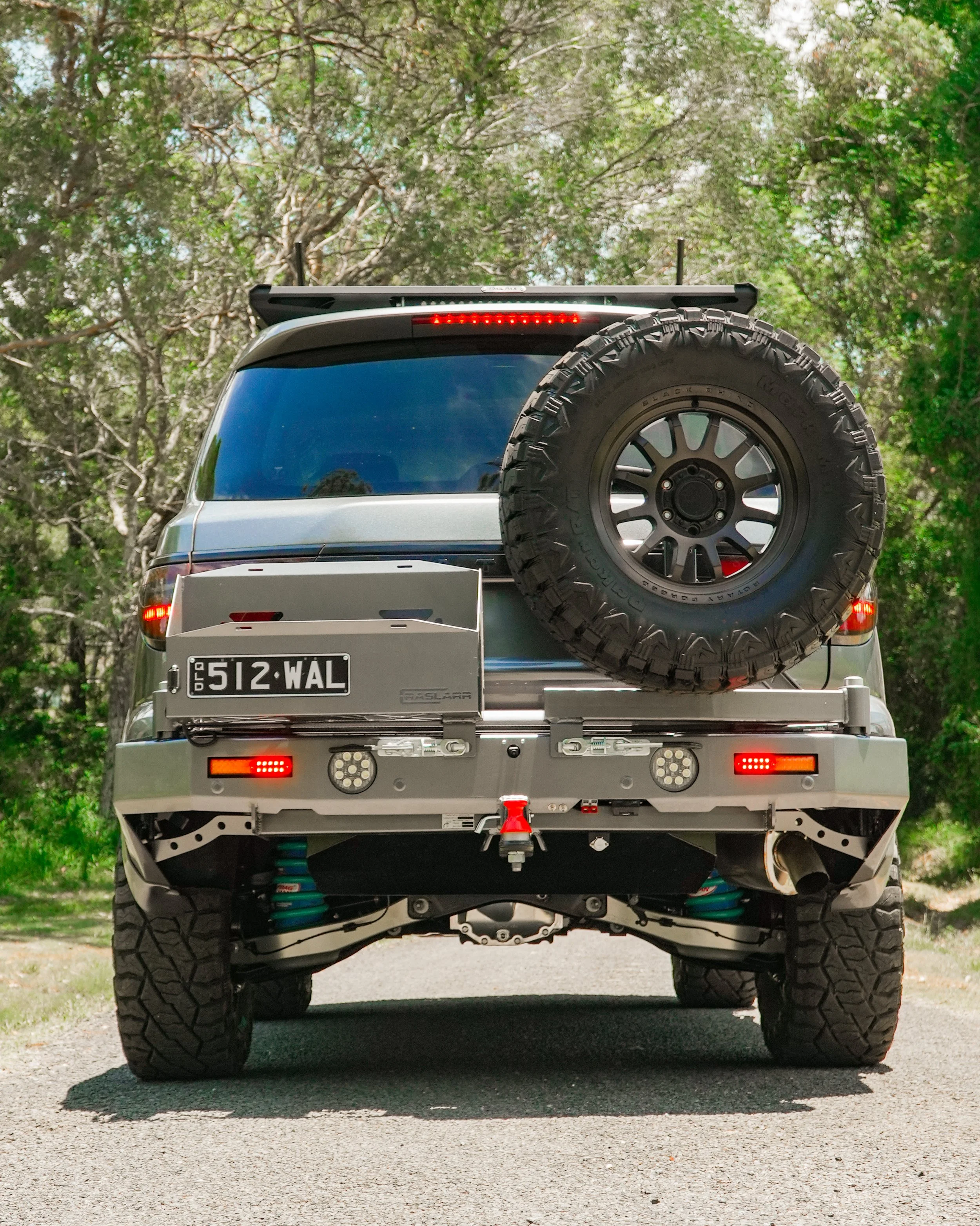 Rear view of a modified off-road vehicle with large tires, a spare tire mounted on the back, and a visible license plate reading '512-WAL,' parked on a paved road with green trees in the background.