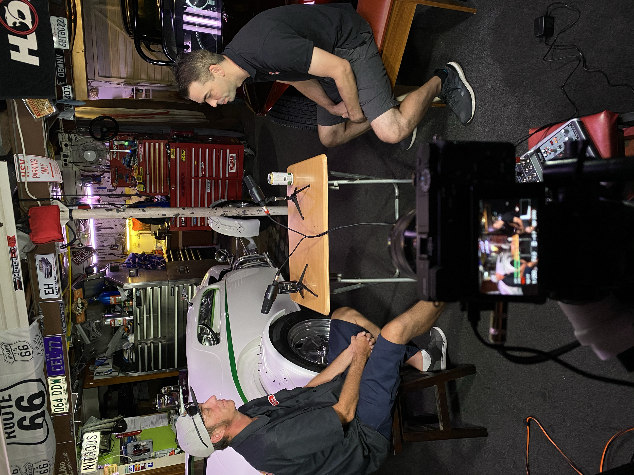 Two men are seated across from each other at a table inside a garage or workshop, with one man working on a small racing car. The garage is decorated with vintage signs and shelves filled with tools and memorabilia.