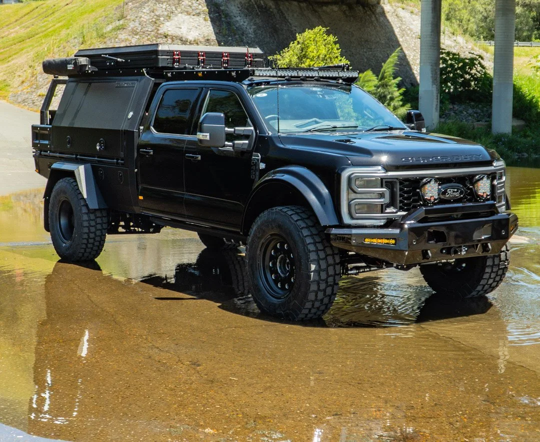 A black Ford Super Duty pickup truck with off-road tires and equipment, parked in shallow water near a creek with greenery and a rocky hill in the background.