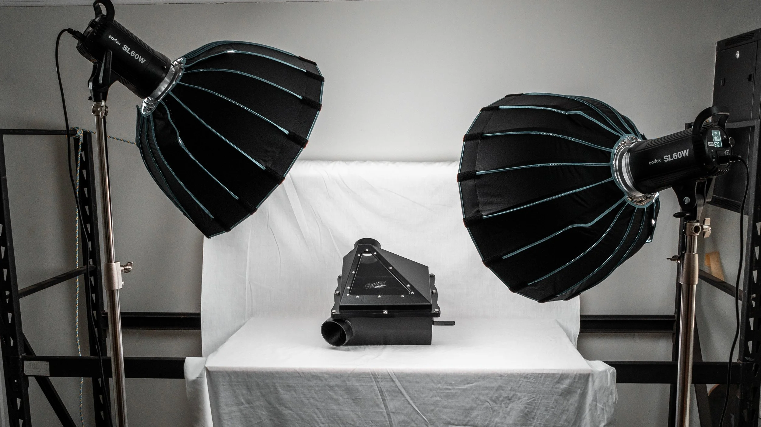 Two large studio strobe lights with black and blue reflectors directed towards a white backdrop, positioned on metal stands on either side of a photography setup. A black digital device is placed on a black stand in front of the backdrop.