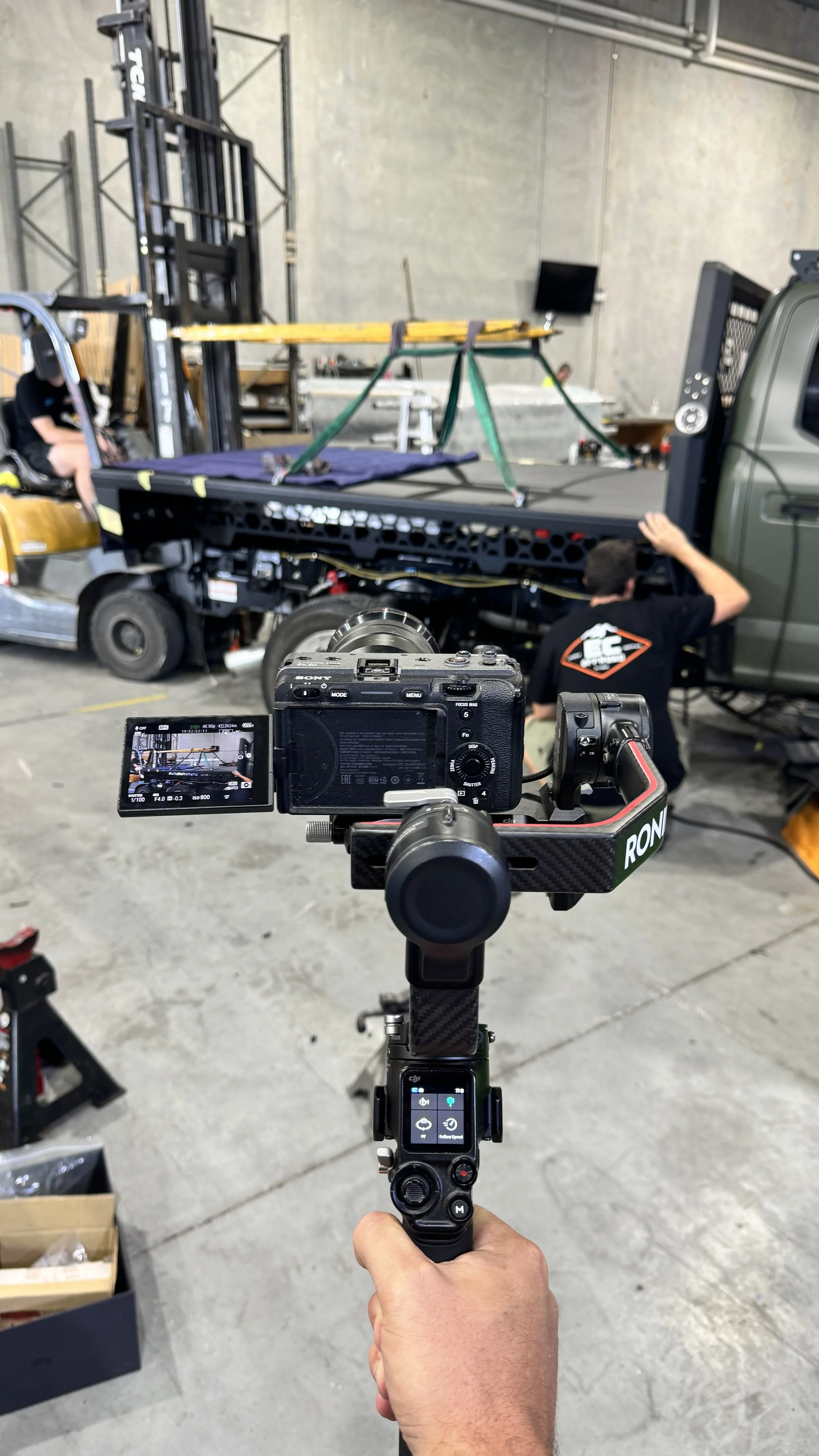 A camera on a stabilizer captures a scene where a person is working on a platform truck, with another person sitting on the truck, in an industrial space.