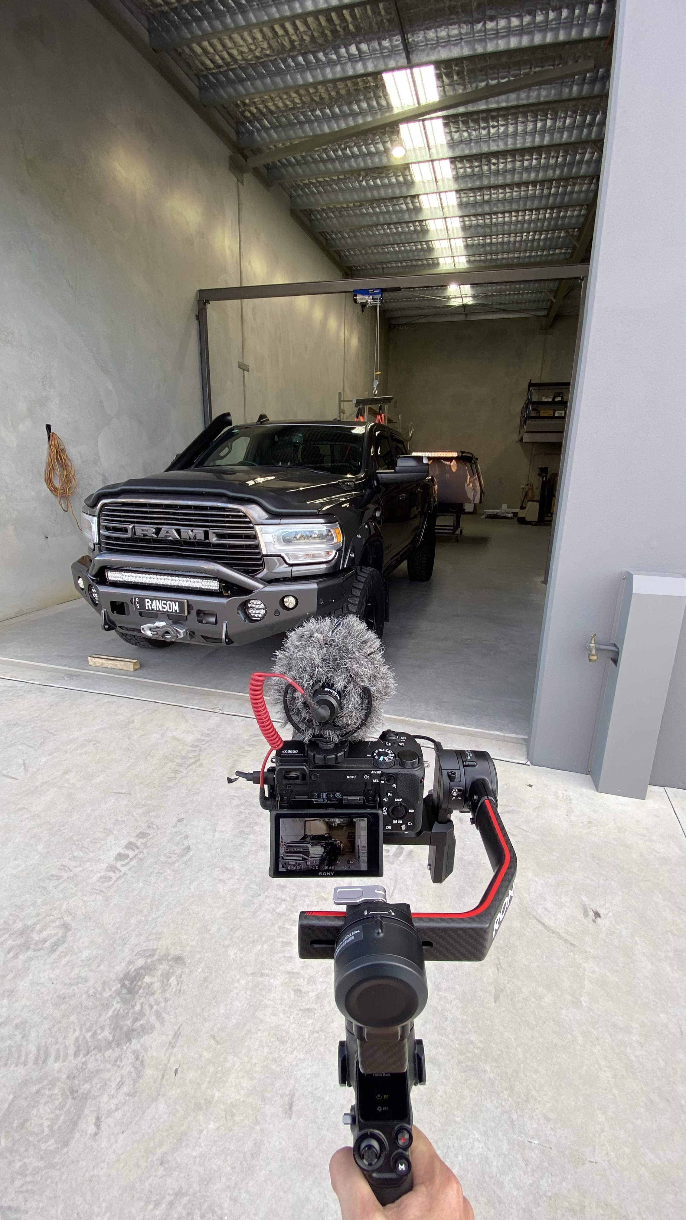 Inside a garage or workshop, a black RAM truck is parked. There is professional camera equipment in the foreground, pointed at the truck, with a microphone attached to the camera. The garage has concrete floors, a high ceiling with metal roofing, and