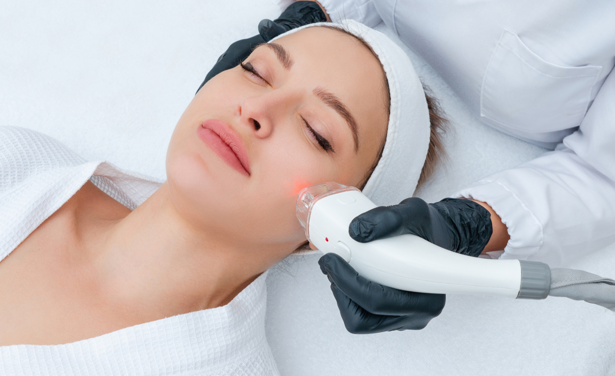 A woman with a headband lying down with eyes closed, receiving a facial treatment with a handheld device emitting red light from a medical professional wearing black gloves.
