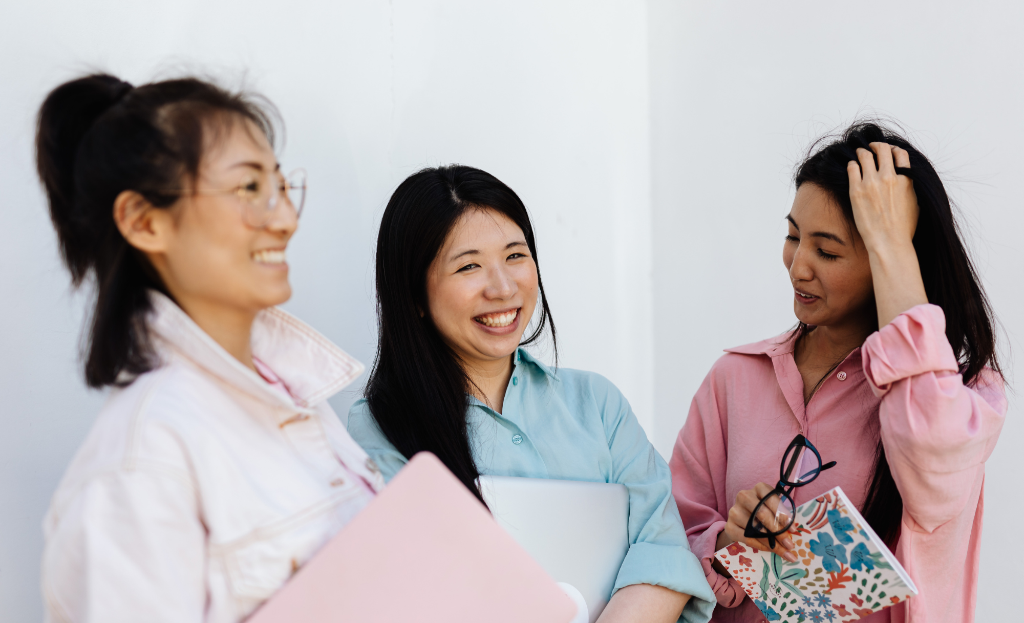 Three women talking and smiling, holding notebooks and a laptop, in an indoor setting.