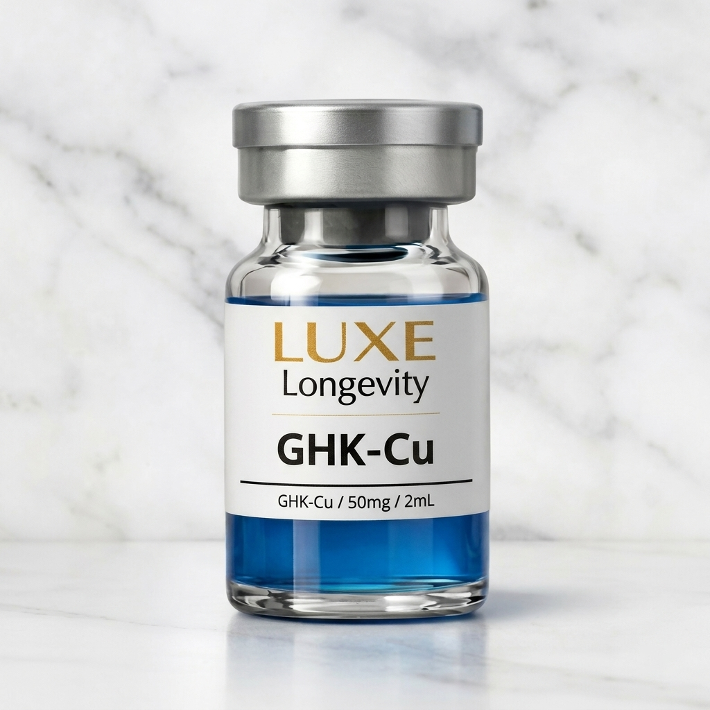 A glass vial labeled 'LUXE Longevity GHK-Cu' with a silver cap, containing a blue liquid, placed on a white surface against a marble background.