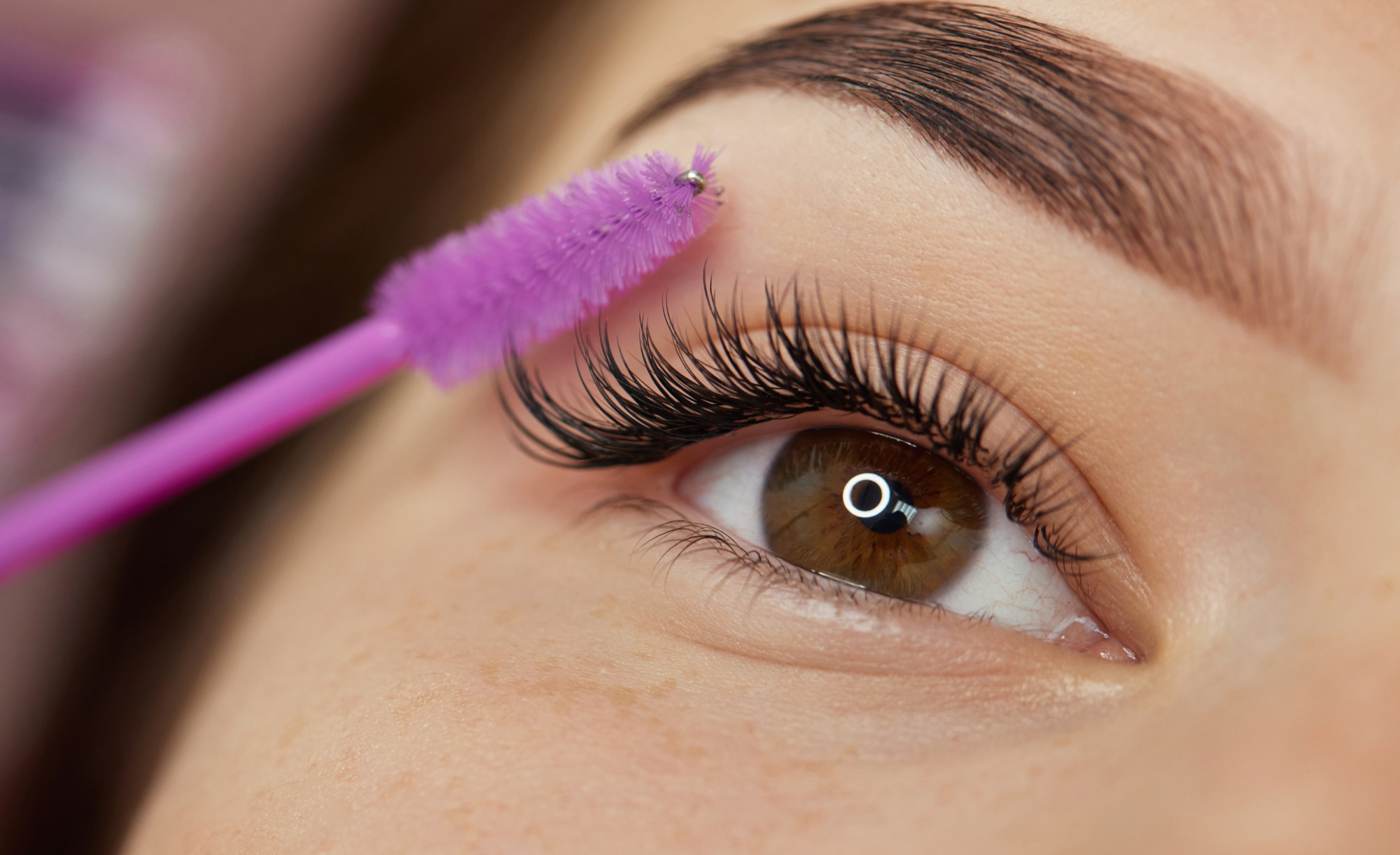 Close-up of a woman's eye with long eyelashes, applying pink eyelash extension glue with a pink applicator.
