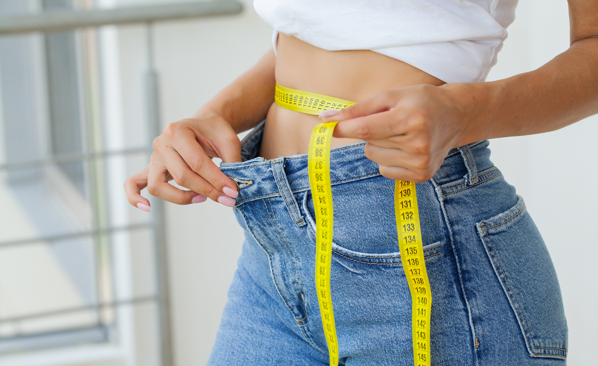 Person measuring their waist with a yellow tape measure, wearing blue jeans and a white top.