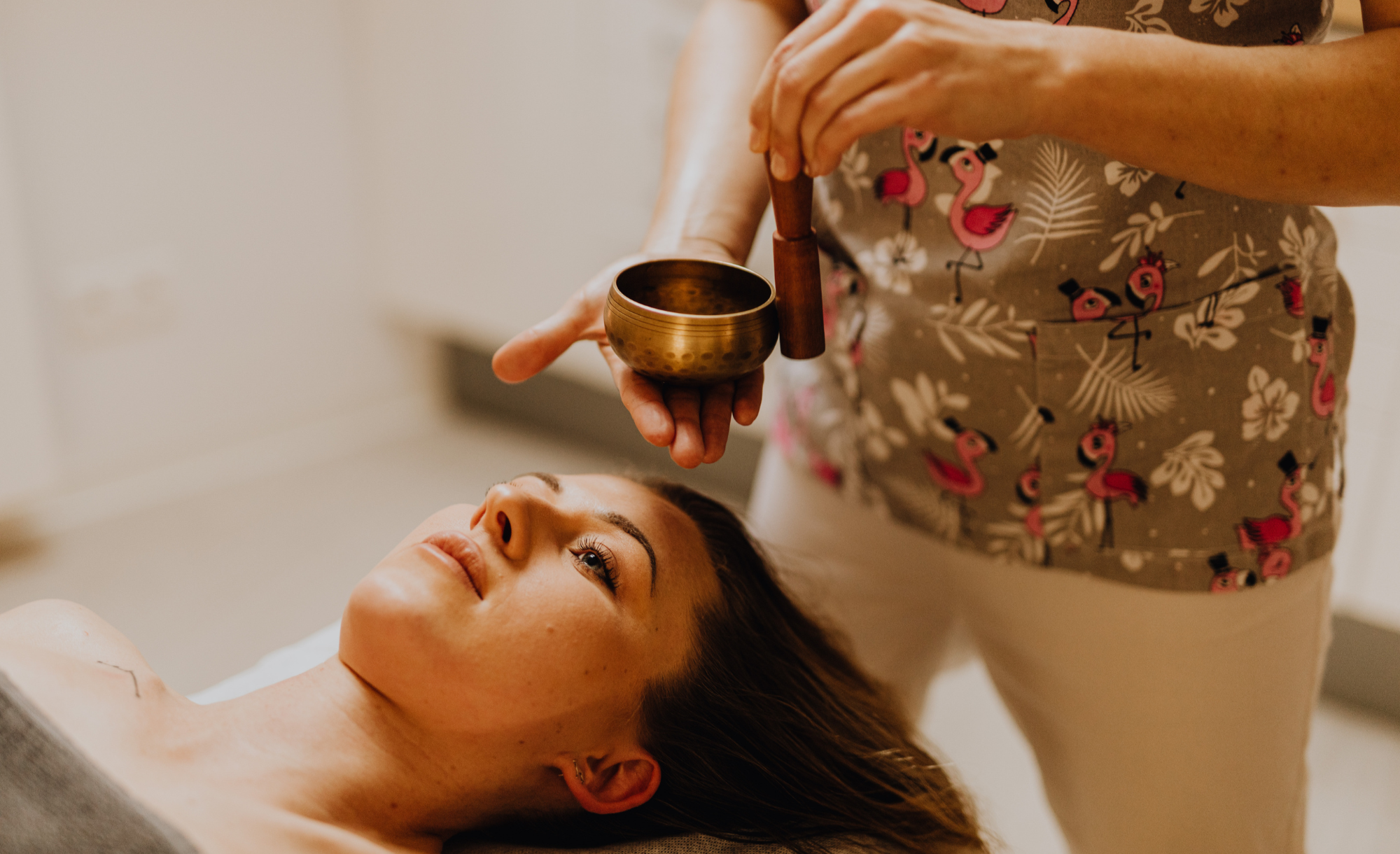 A woman lying on a massage table receiving sound therapy with a singing bowl and mallet from a therapist wearing a flamingo patterned shirt.