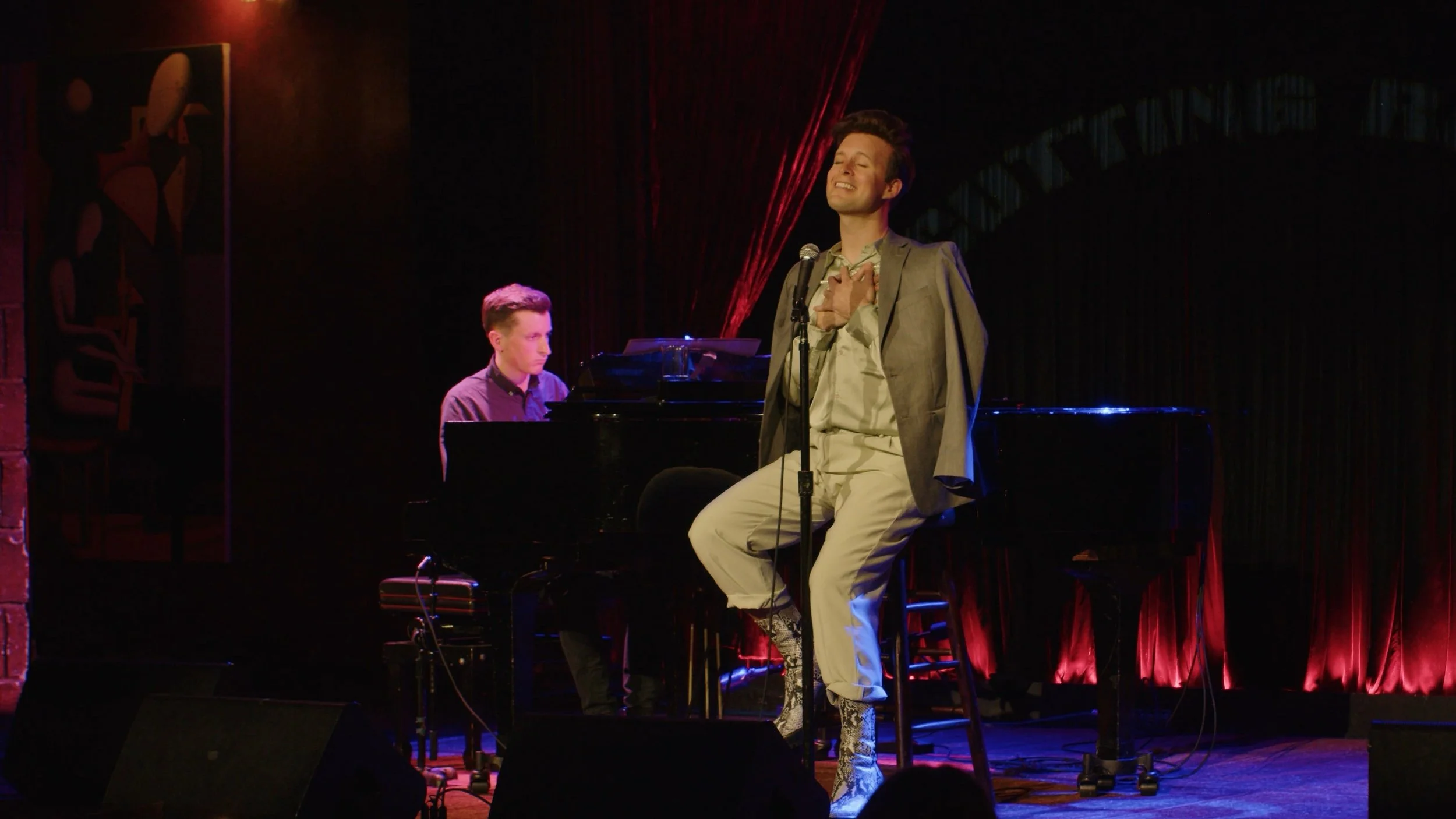 Man sits on a chair and sings into a microphone. There is an accompanist in the background.