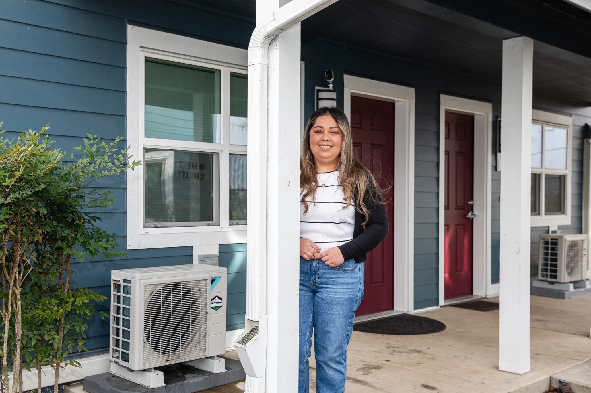 A smiling woman in casual clothing standing on a porch of a blue house, with windows, a red door, and air conditioning units visible.
