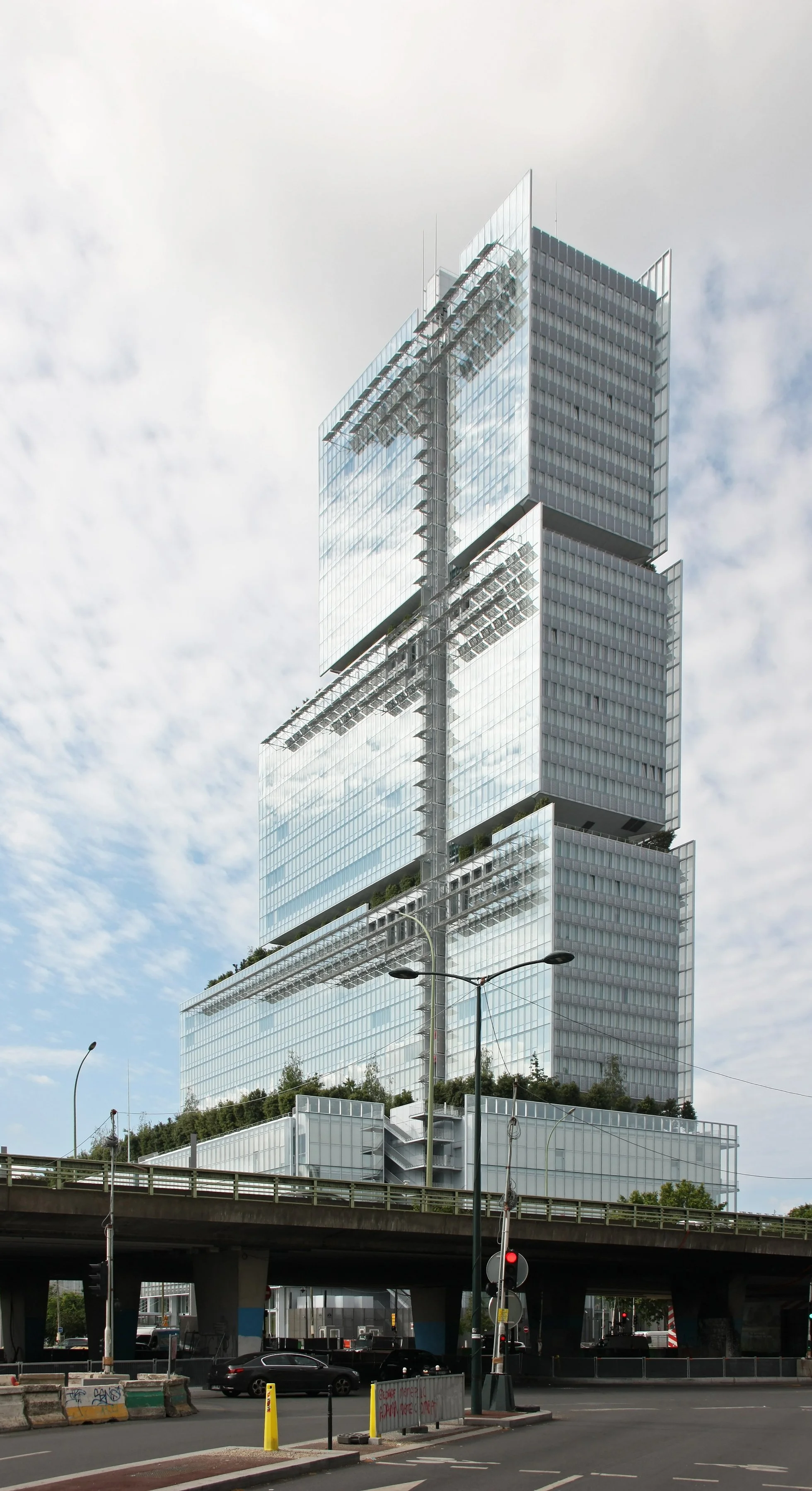 Gratte-ciel moderne avec plusieurs niveaux et façades en verre, entouré de lampadaires et de véhicules, avec un ciel partiellement nuageux.