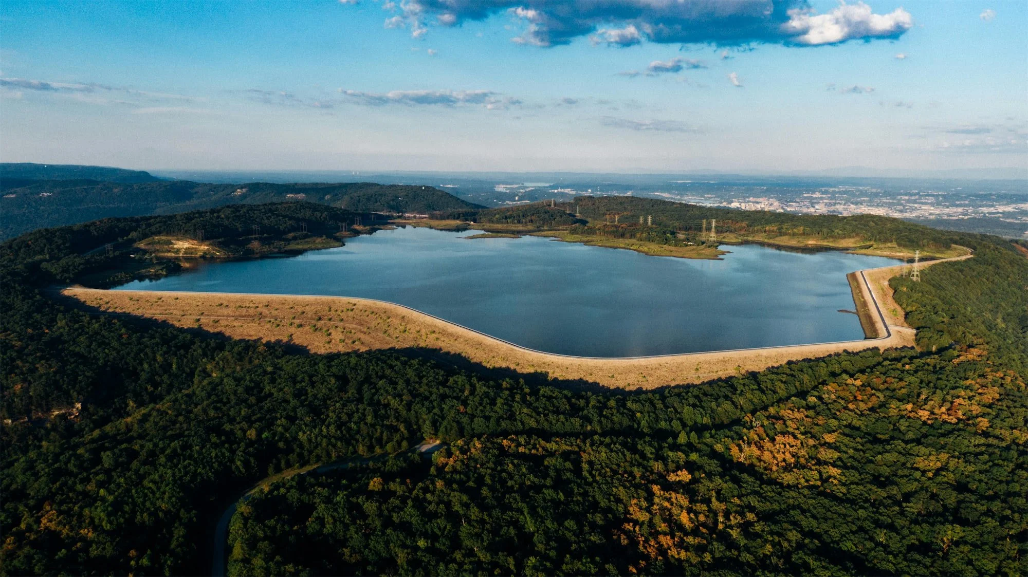 An aerial view of a large reservoir surrounded by green forested hills with a dam on one side, under a partly cloudy sky.