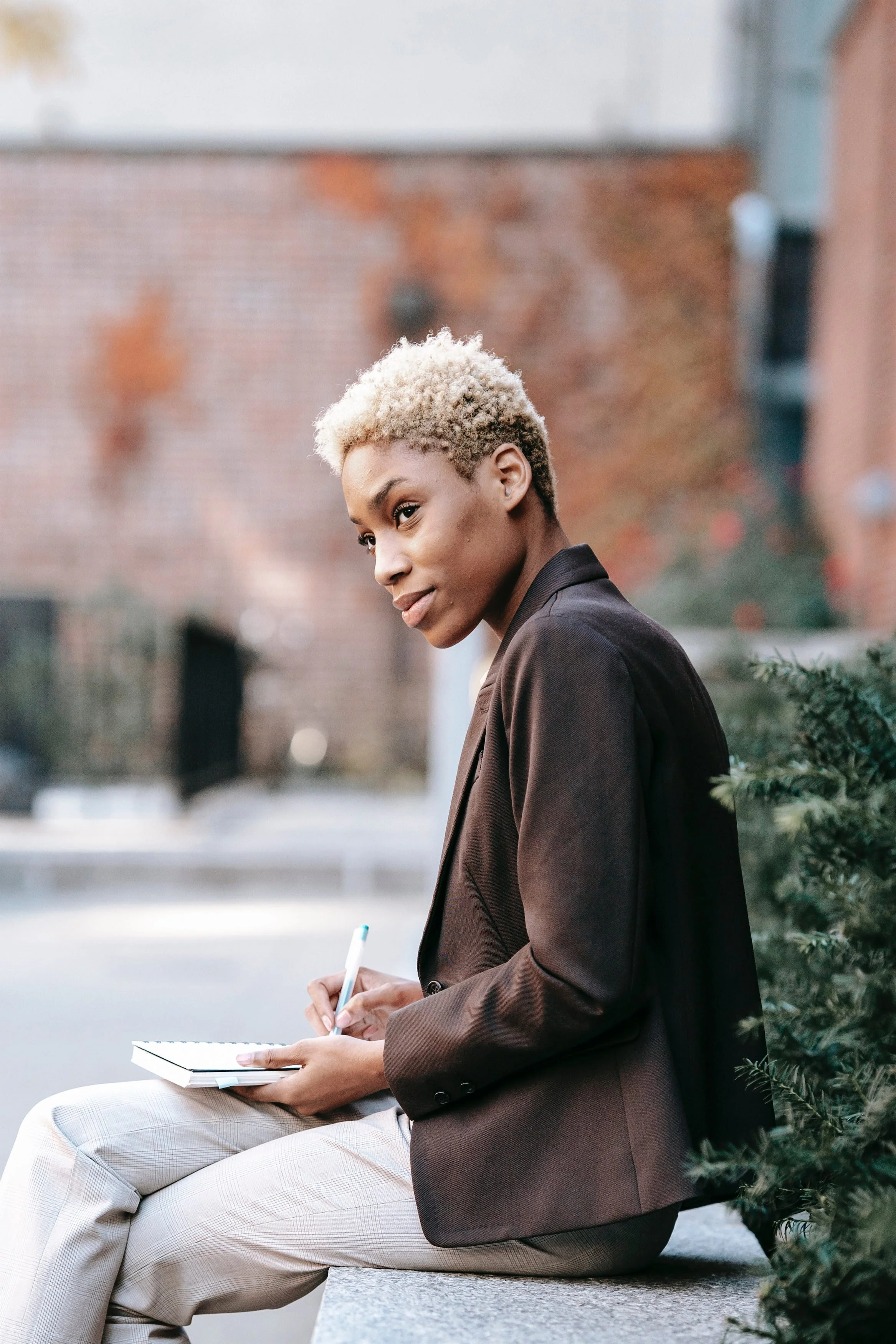 Young woman with blonde curly hair sitting on a bench outdoors, writing in a notebook and smiling at the camera, with blurred background of brick wall and urban environment.