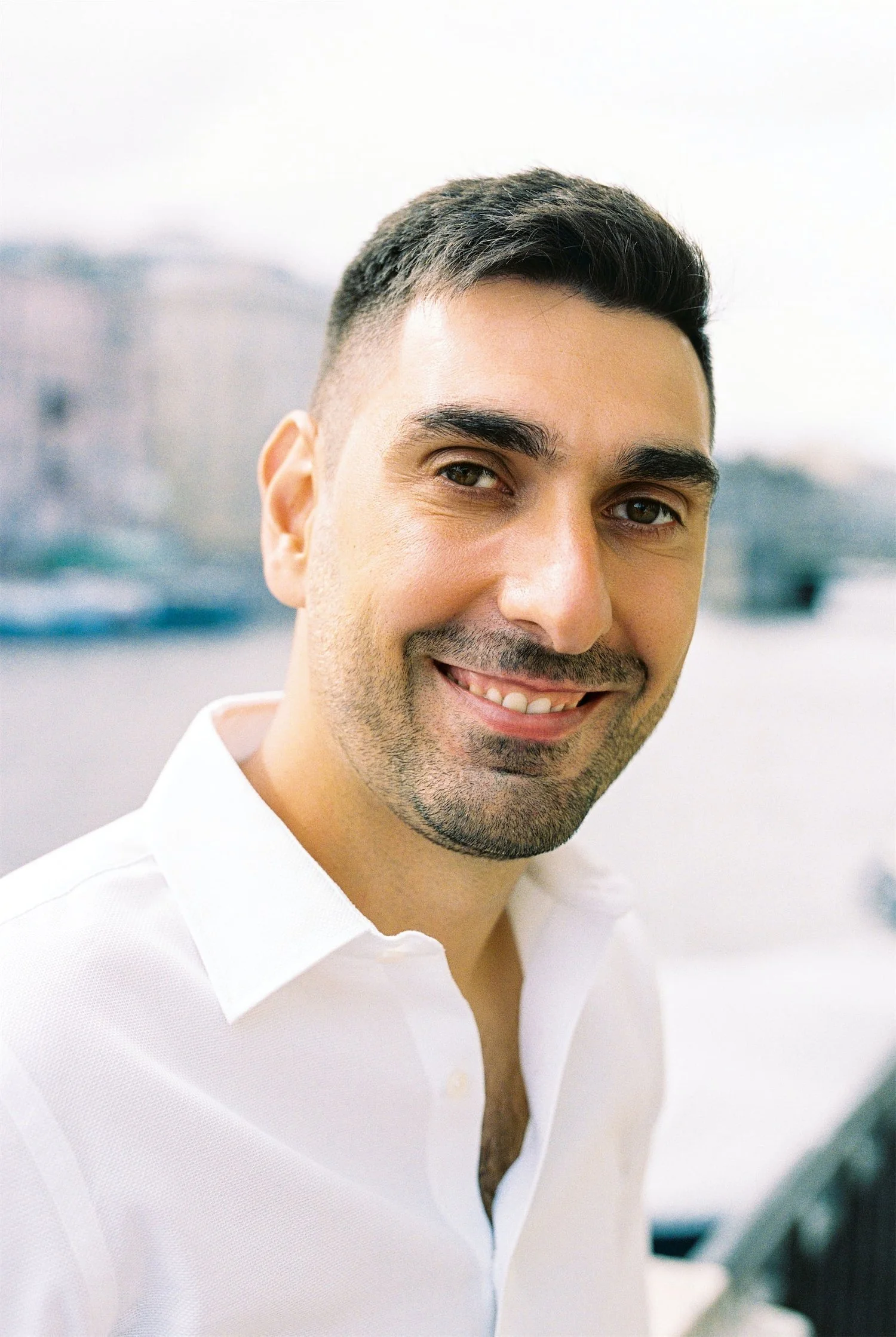 Close-up of a smiling man with short dark hair, wearing a white shirt, outdoors with blurred background.