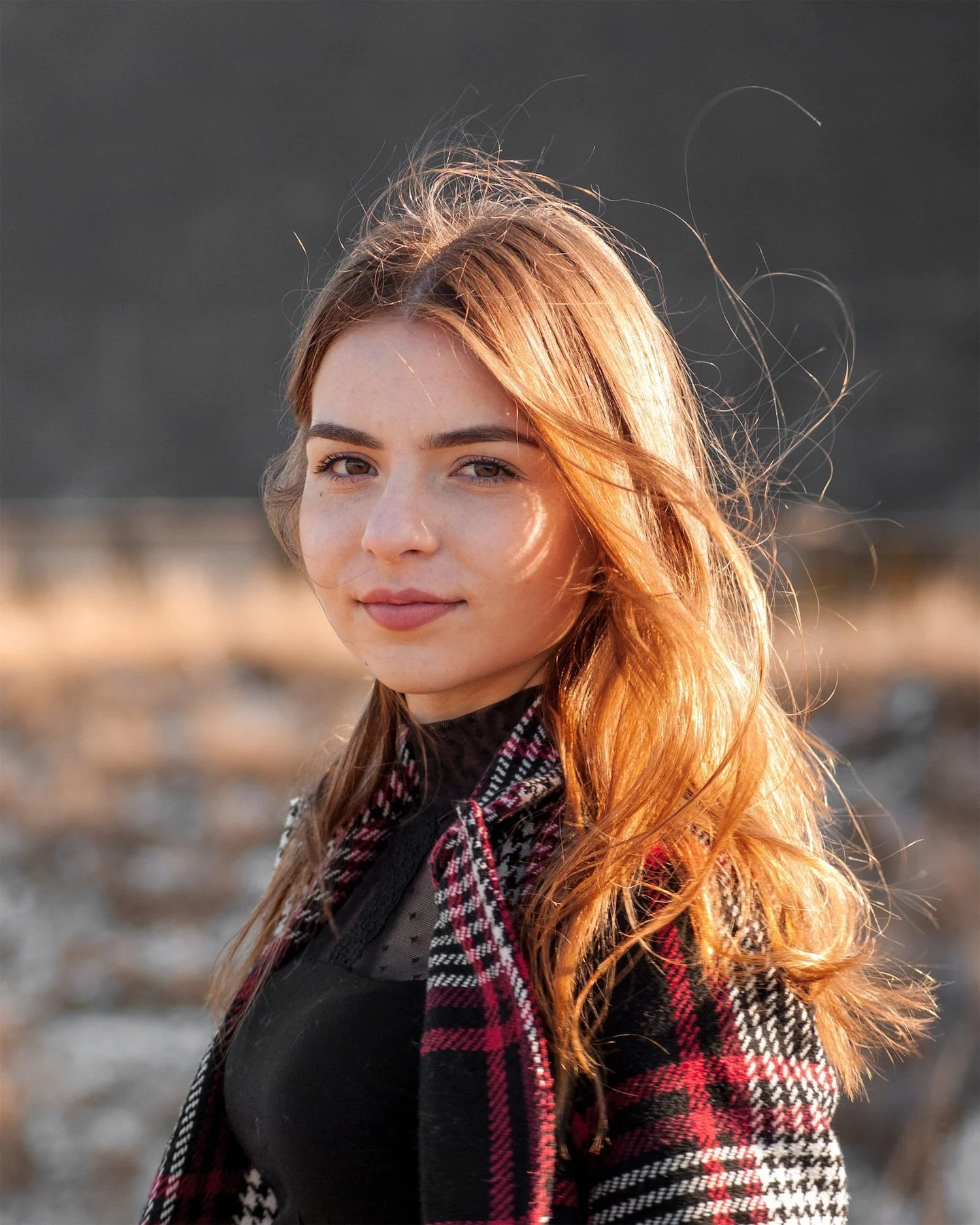 A young woman with long red hair smiling outdoors in sunlight, wearing a black top and a red and black plaid coat.