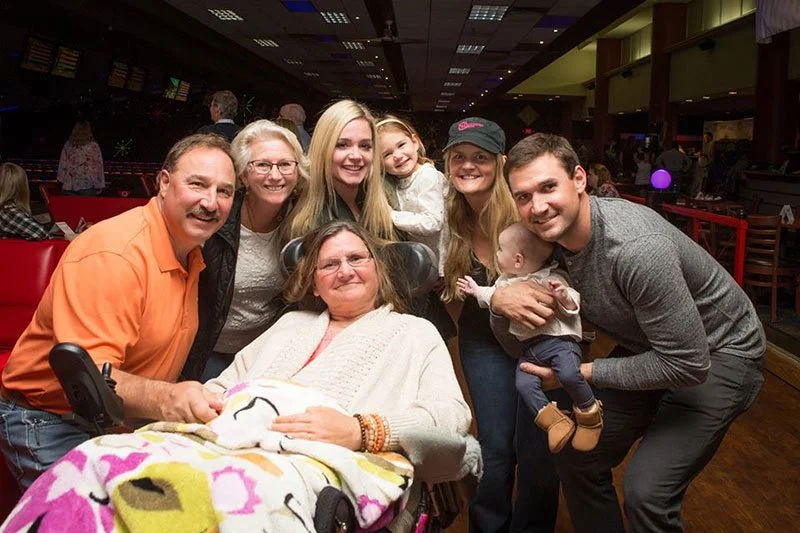 A group of seven people, including an elderly woman in a wheelchair, smiling and posing for a photo in an indoor venue with a dark ceiling and socializing in the background.
