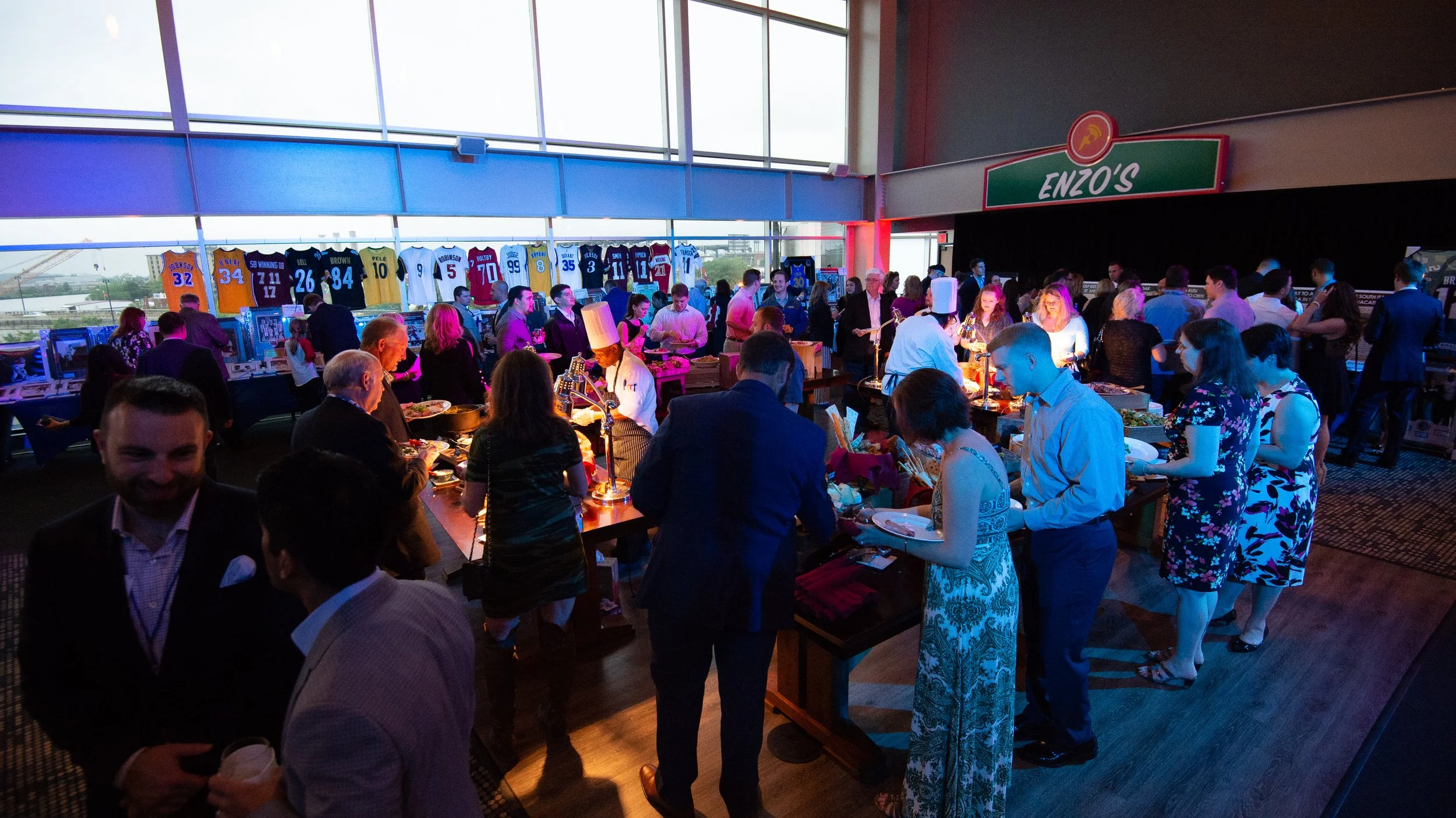 Indoor event at a sports stadium or arena featuring a lively crowd, with framed sports jerseys displayed on a high wall and tables where people are eating and socializing.