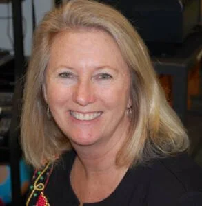 A smiling woman with blonde hair, wearing earrings and a black top, in an indoor setting.