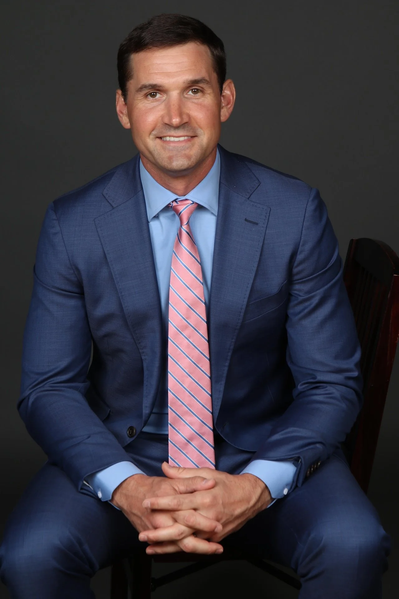 MLB Athlete Ryan Zimmerman in a blue suit and pink striped tie sitting on a dark wooden chair against a plain dark background.