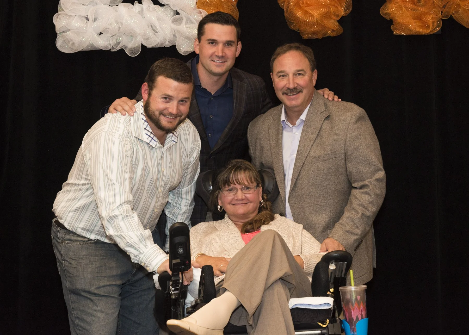 Group of six people, including a woman in a wheelchair, posing together in front of a black background with orange and white decorative decorations. They are smiling, with some resting hands on each other's shoulders.