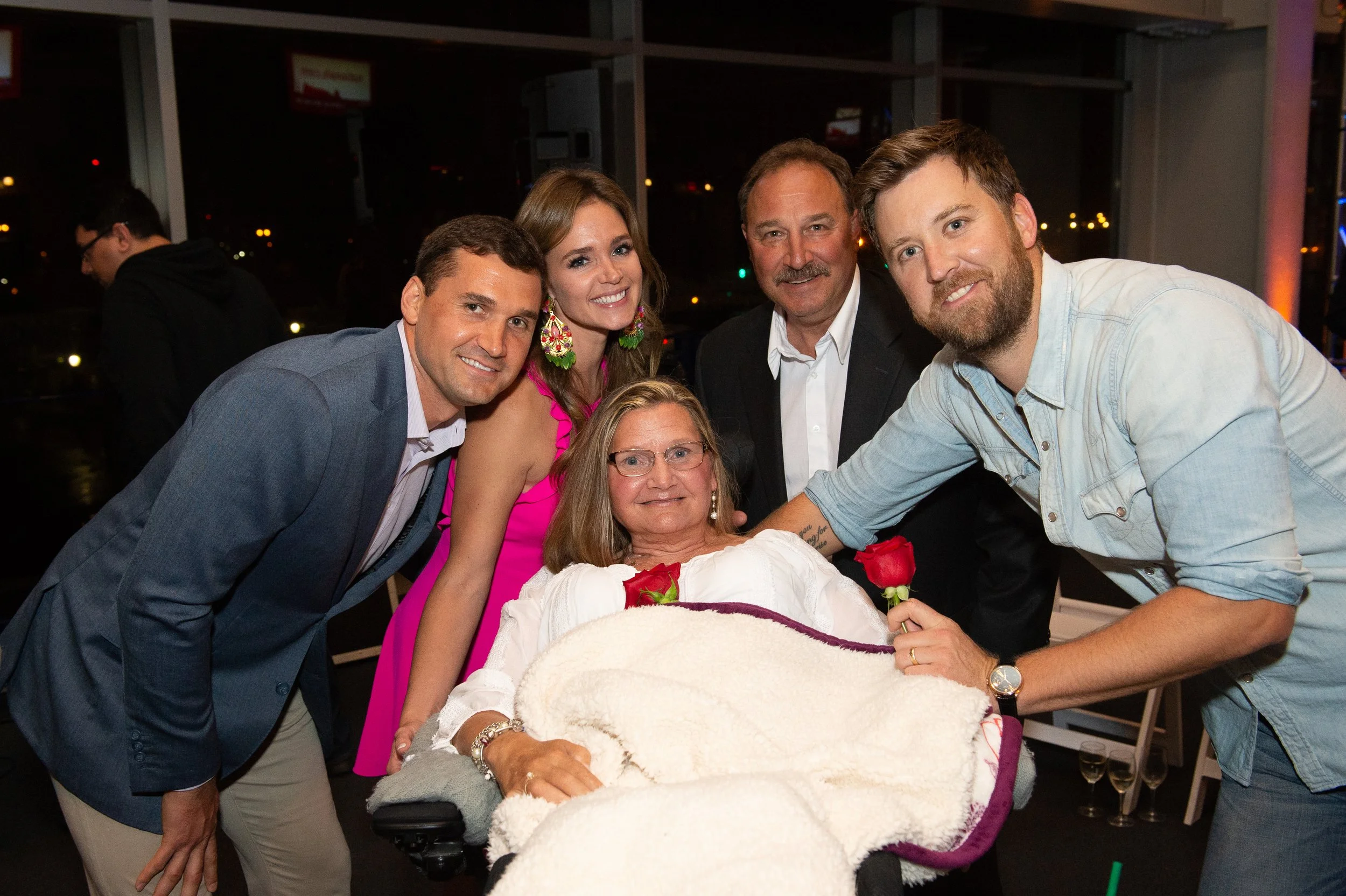A group of six people gathered around a woman in a wheelchair, holding a red rose, with smiling faces at a social event or celebration.
