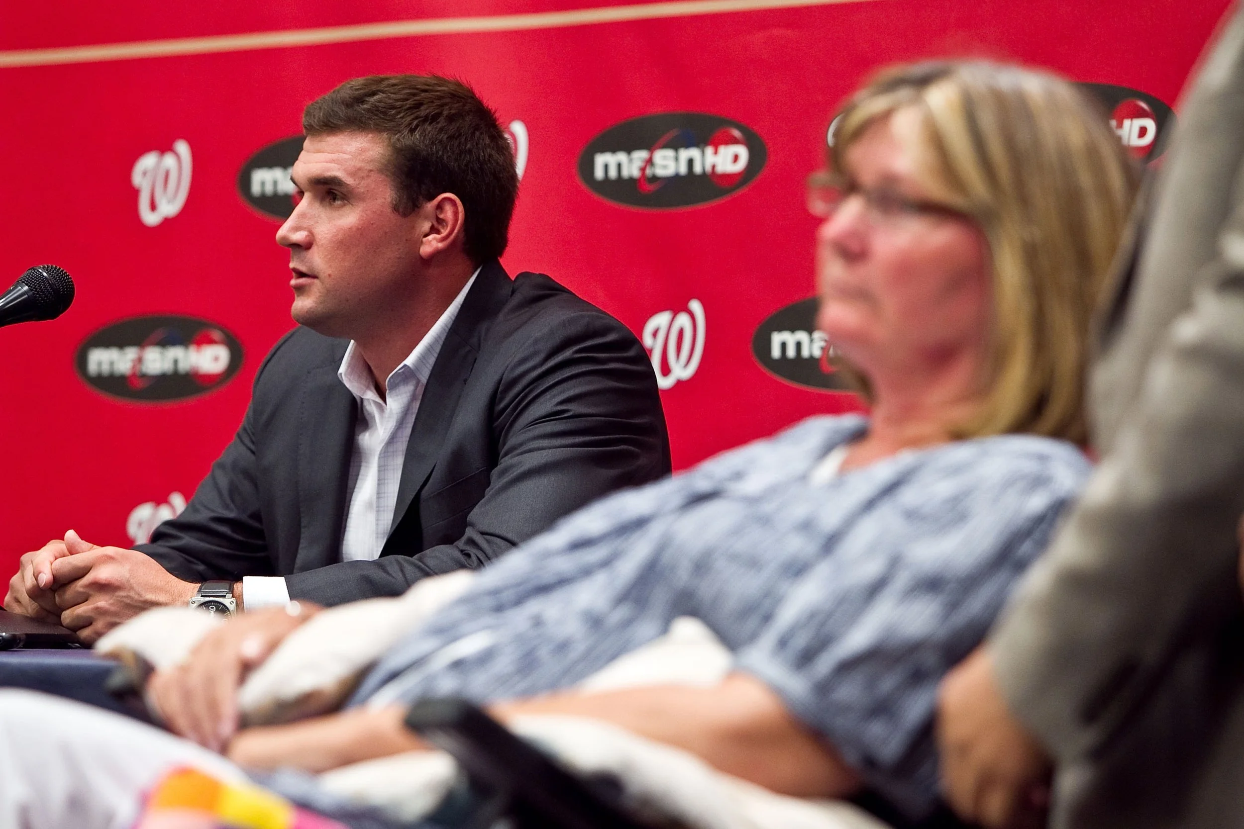 A man in a suit seated at a press conference table with a microphone, with a woman sitting next to him, blurred in the foreground, both against a red background with logos.