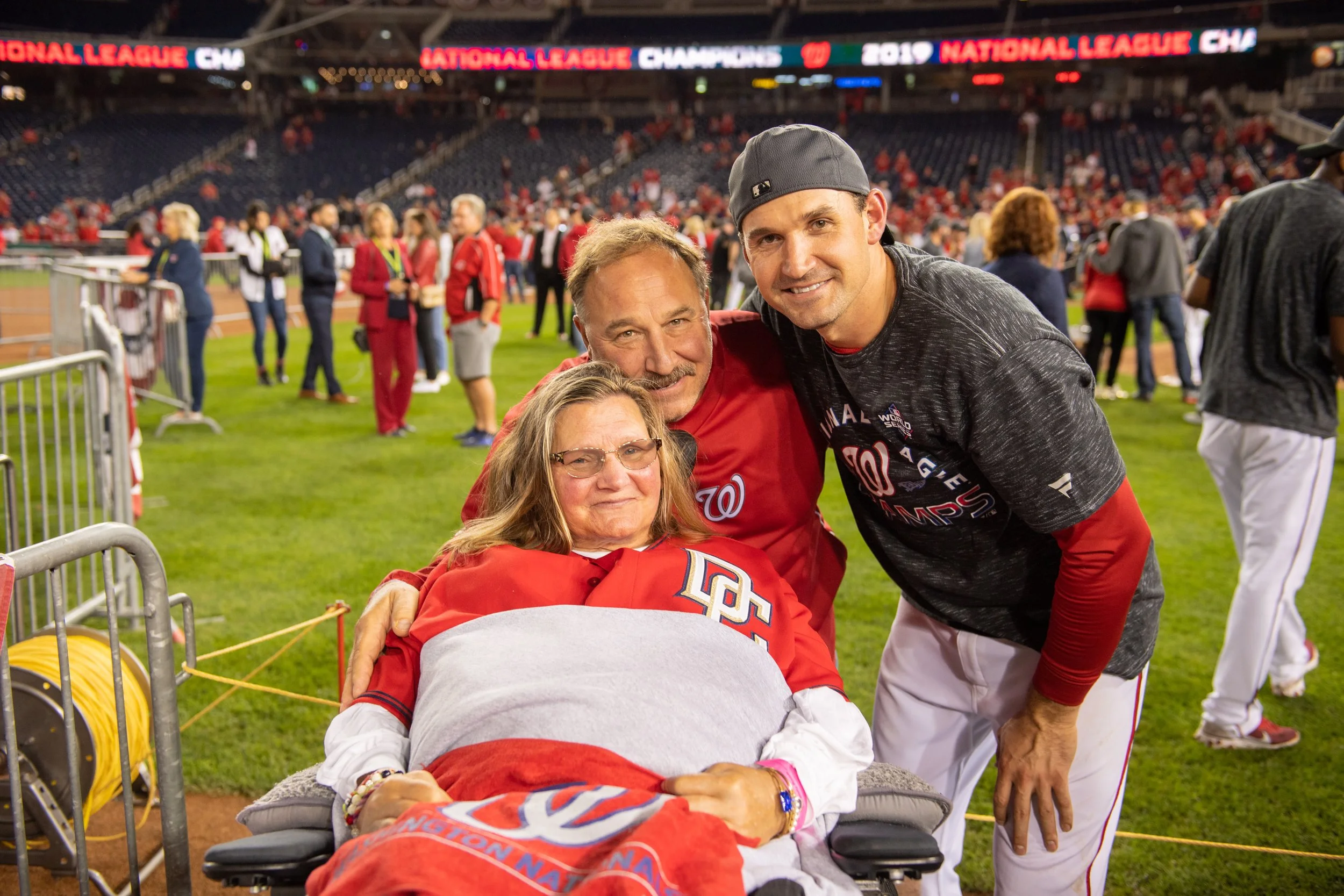 Three people celebrating at a baseball game, with one person in a wheelchair and wearing a Washington Nationals jersey, two others smiling, all in a stadium with many other spectators and a digital banner reading "National League Champions 2019."