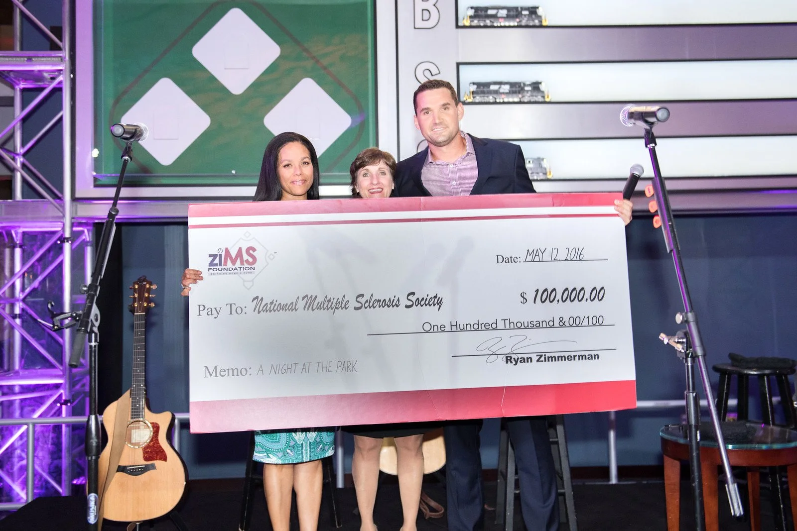 Three women and one man on a stage holding an oversized check for $100,000 made out to the National Multiple Sclerosis Society, dated May 12, 2016, with a memo 'A Night at the Park'. There are microphones, a guitar, and stage equipment visible, with 