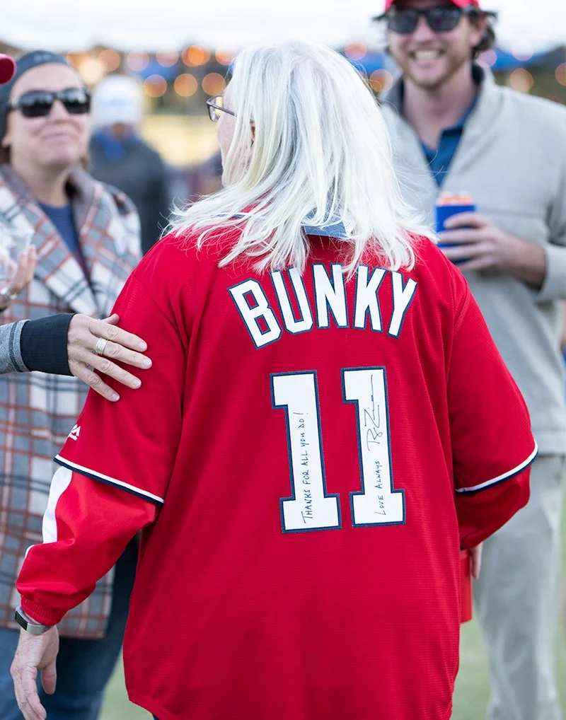 A woman with blonde hair wearing a red jersey with the name BUNKY and the number 11 on the back, signing messages on her jersey. She is surrounded by people at an outdoor event.