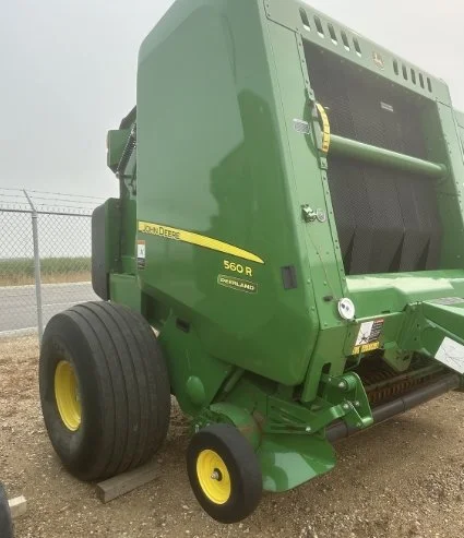 Green John Deere 560 round baler parked outdoors on a dirt surface, with a fence in the background.