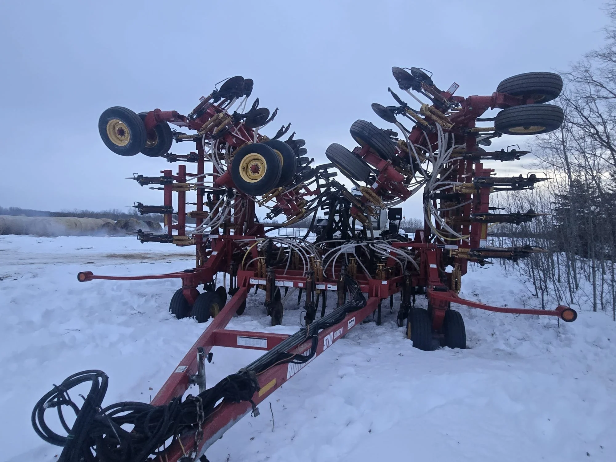 A red farm equipment attachment with multiple wheels and hydraulic hoses outdoors in a snow-covered area, with trees in the background.