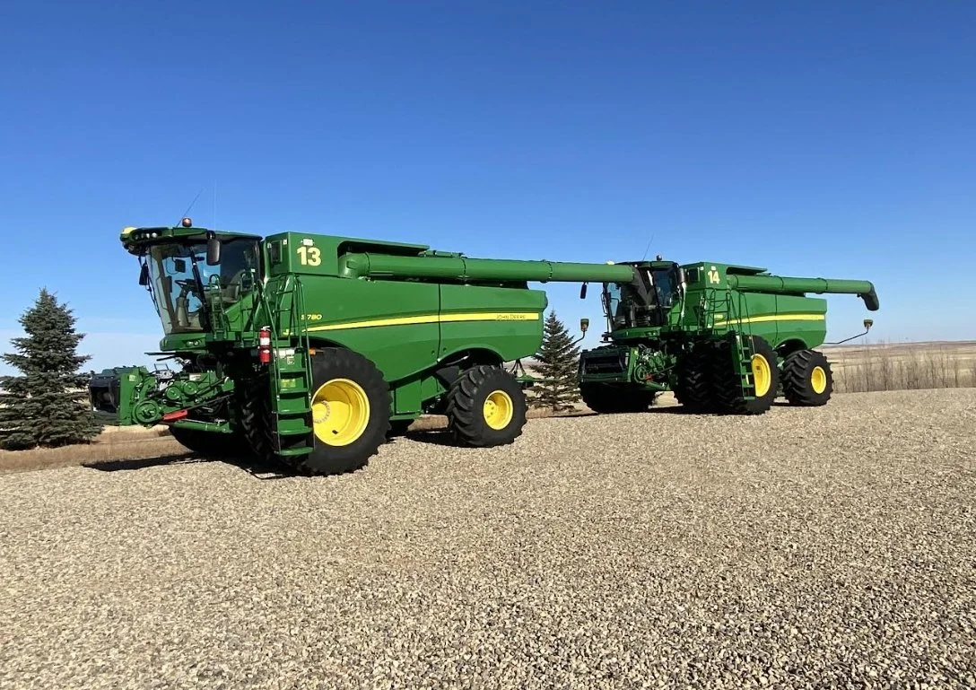 Two large green John Deere combine harvesters parked on gravel under a clear blue sky, with a few small trees and open field in the background.