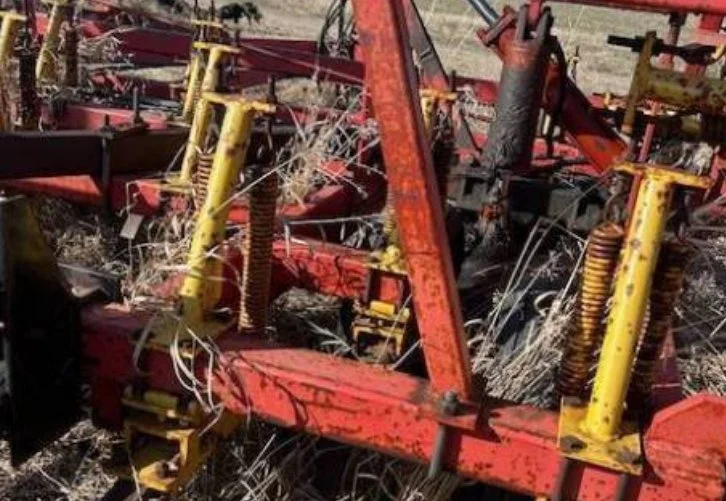 Close-up of rusted, fallen farm equipment with yellow and red parts amid dry grass and weeds in a field.