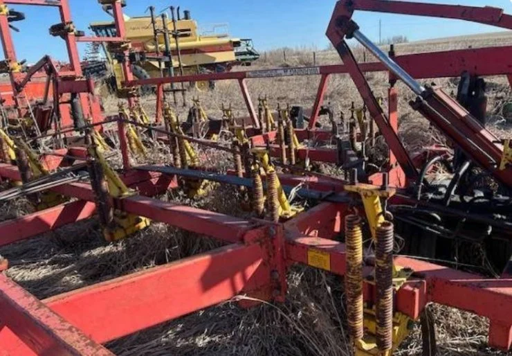 Close-up of a red farming cultivator with yellow springs, set in a field with dry grass, under a clear blue sky.